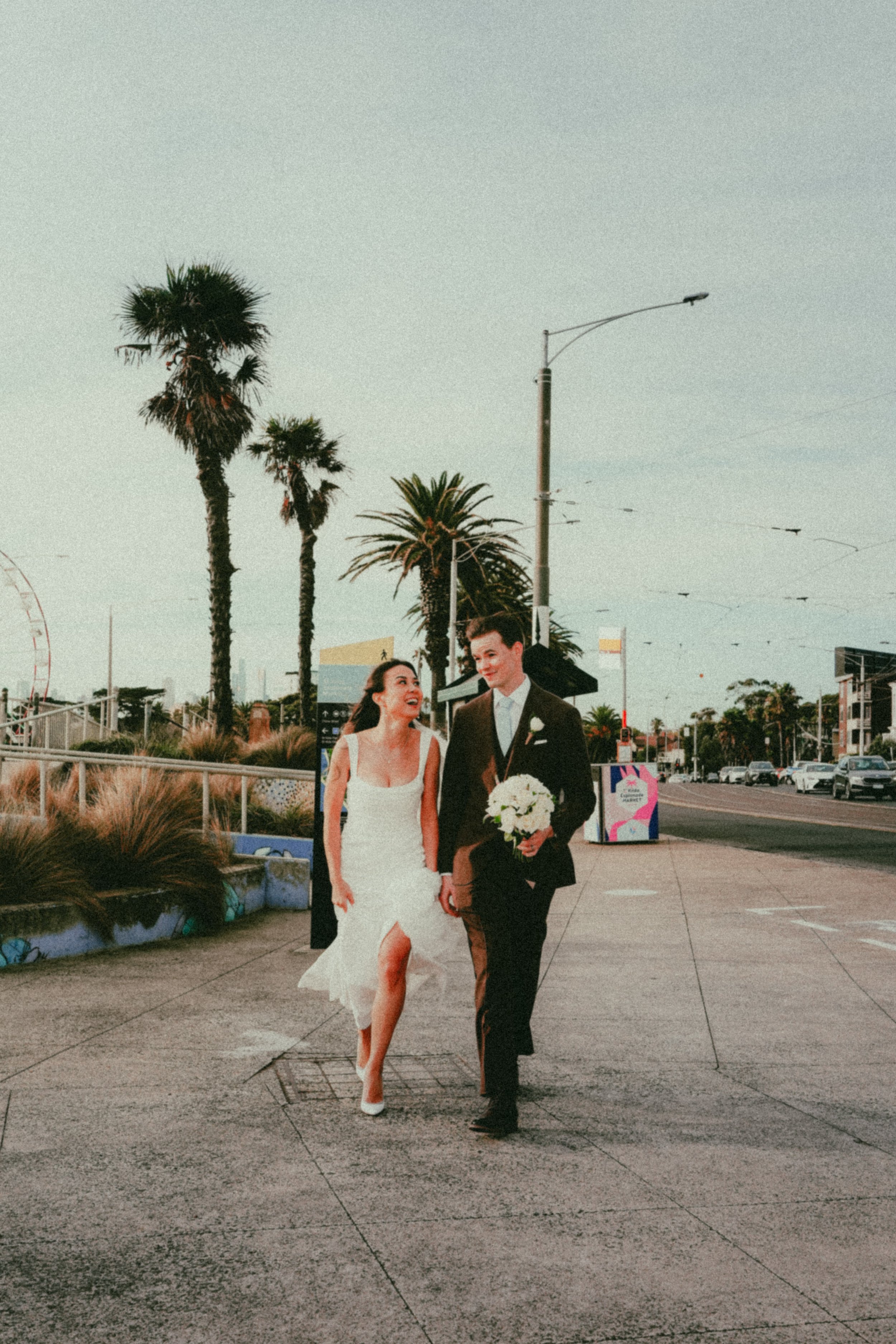 A bride and groom walk hand-in-hand outdoors in a city setting with palm trees and a Ferris wheel in the background, during sunset.