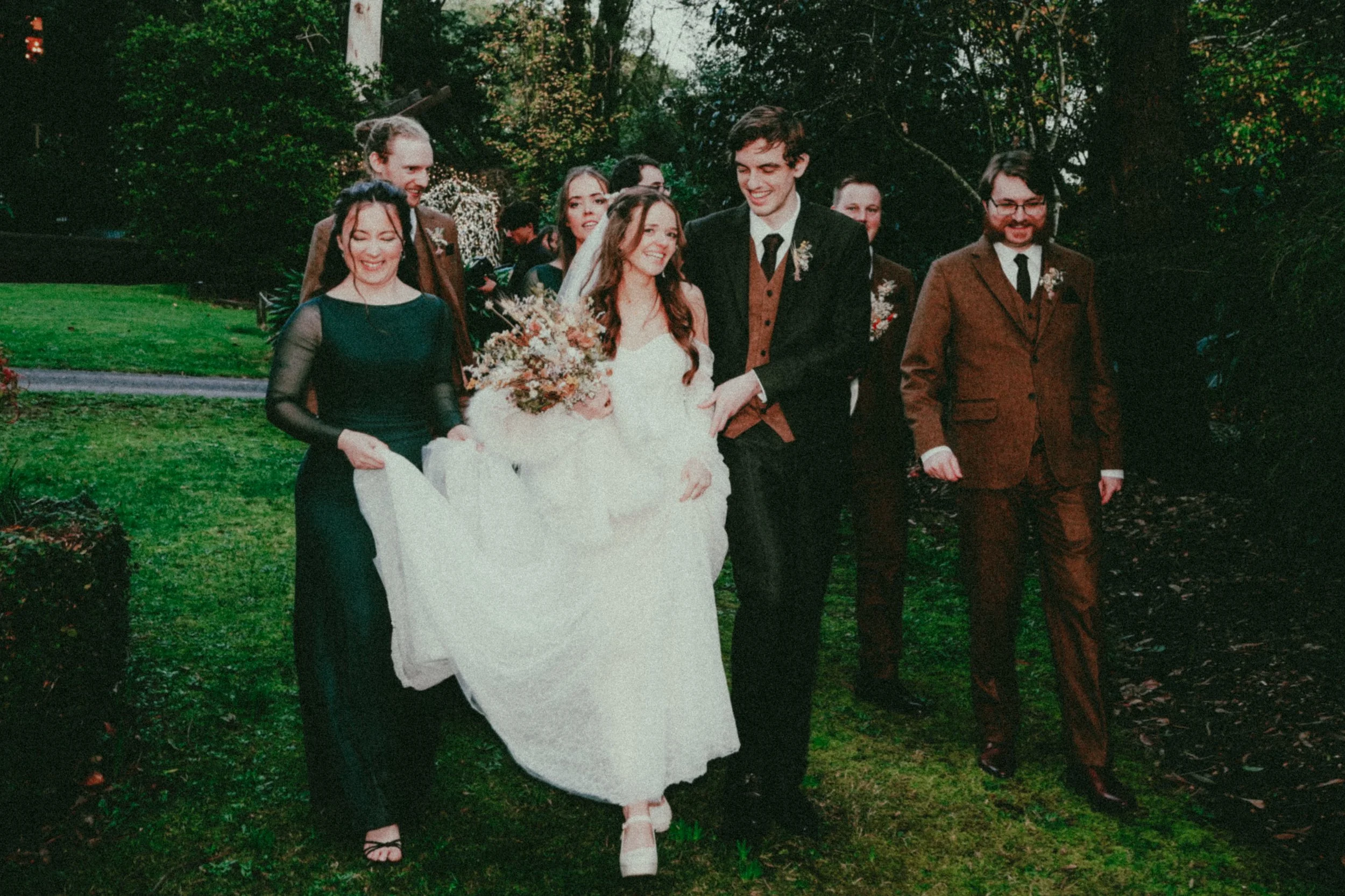 A group of people walking outdoors in a park with trees and grass, celebrating a wedding, with a bride in a white dress and a groom in a dark suit, surrounded by friends.