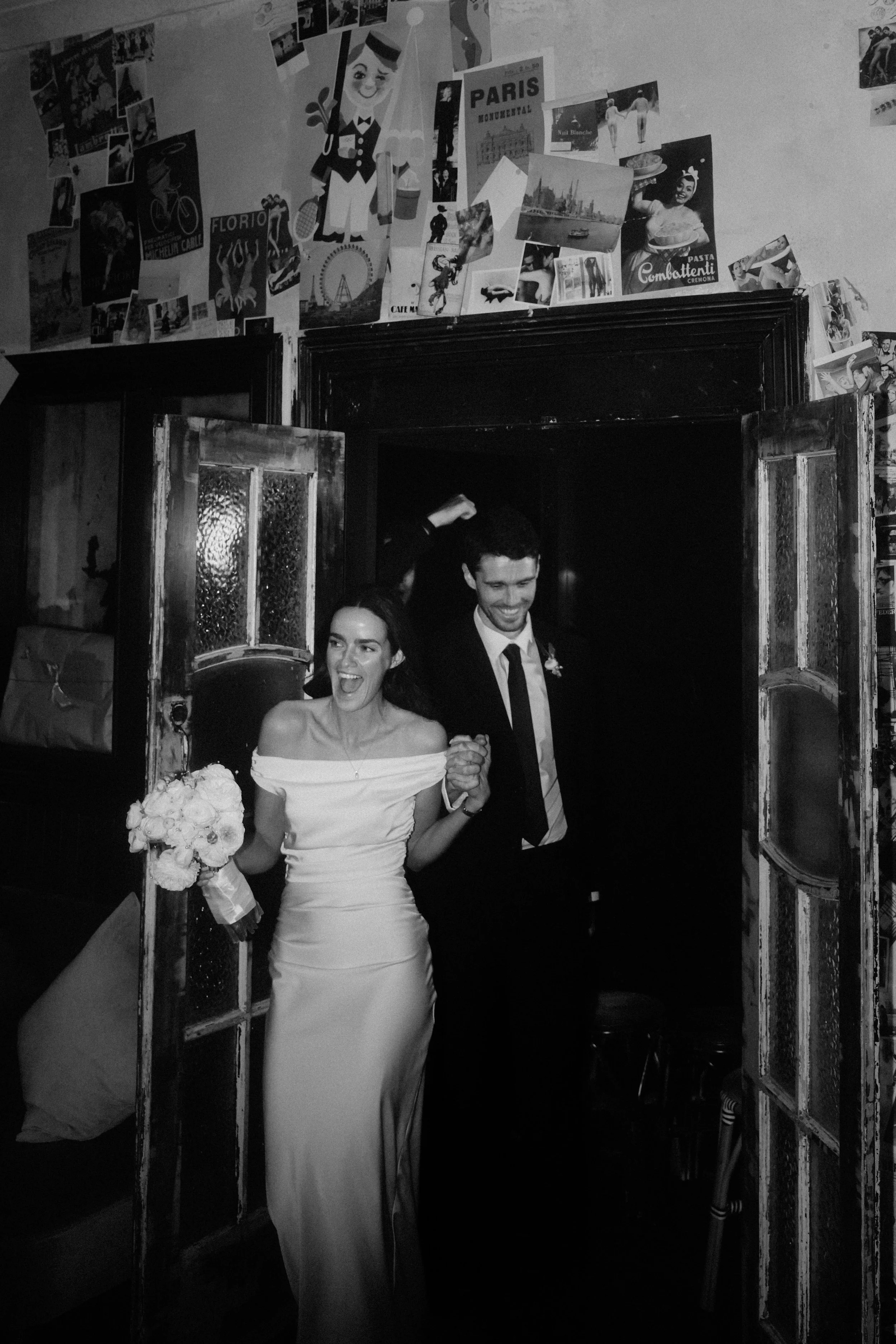 A bride and groom holding hands and smiling as they walk out of a doorway, the bride holding a bouquet of flowers, with a wall decorated with various posters and photographs behind them.