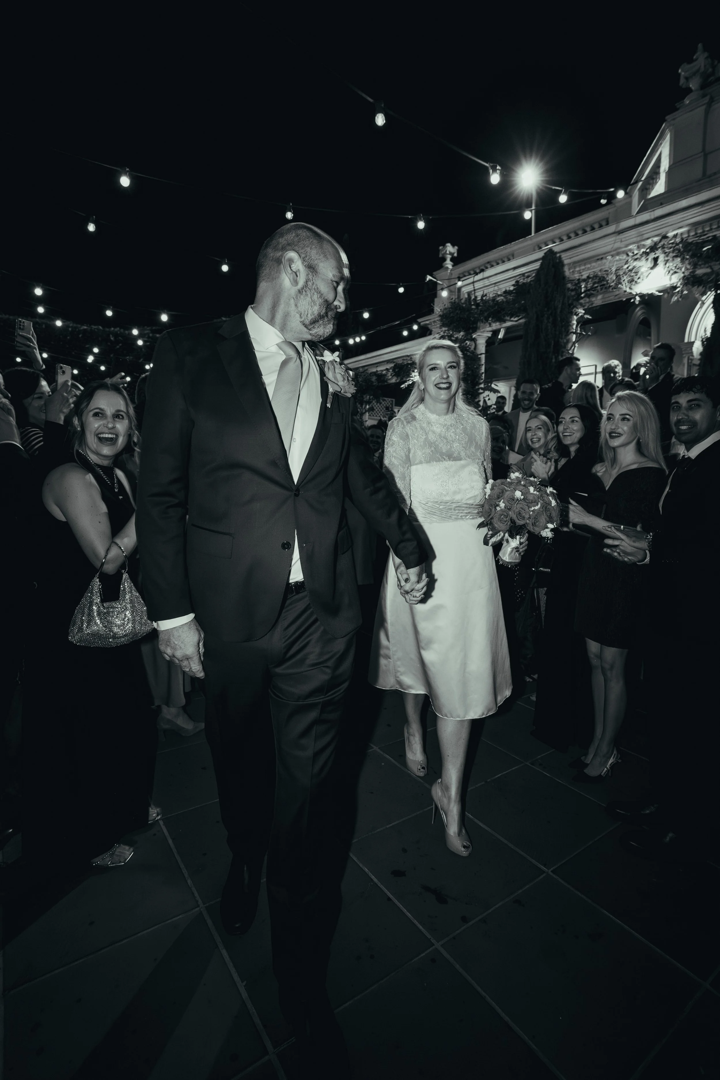 A wedding reception at night with string lights overhead. An older man in a suit and a woman in a wedding dress are holding hands and walking together. The woman is smiling and holding a bouquet. Several guests are smiling, clapping, and taking photo