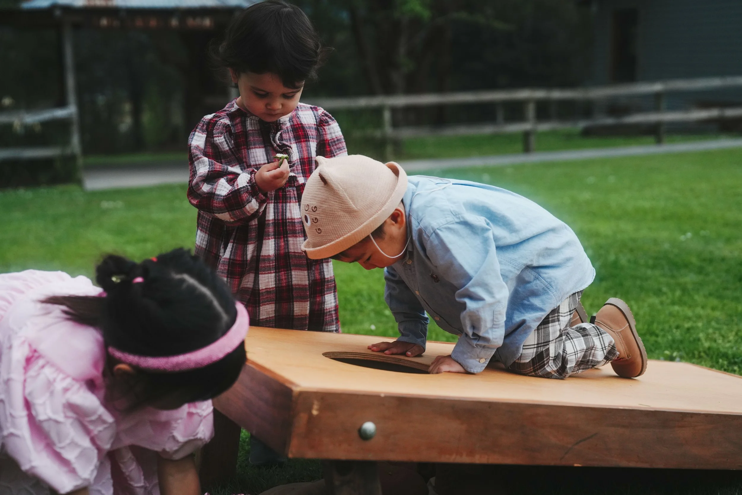Children playing a game on a wooden table outdoors with a grassy background. One girl is standing, holding something in her hand, while a boy is kneeling and leaning over the table. A girl in the foreground, wearing a pink jacket, is observing the ga