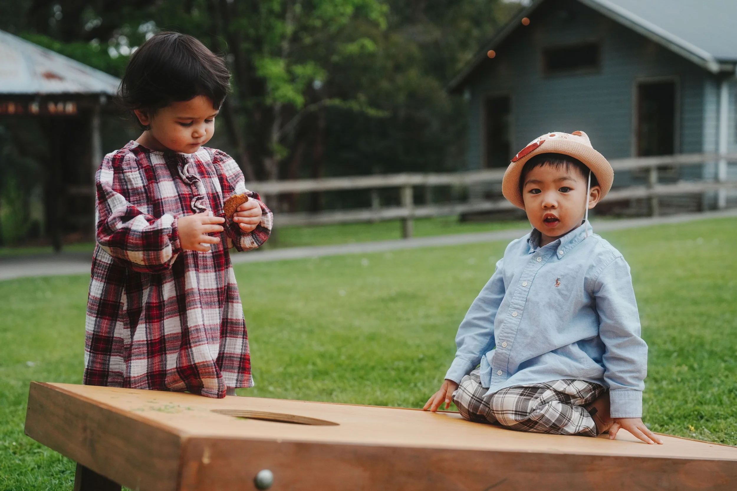 Two children playing buckaroo game outdoors on a wooden board, with one child in a plaid dress and holding a bean bag, and the other child kneeling on the board wearing a hat and checkered shorts.
