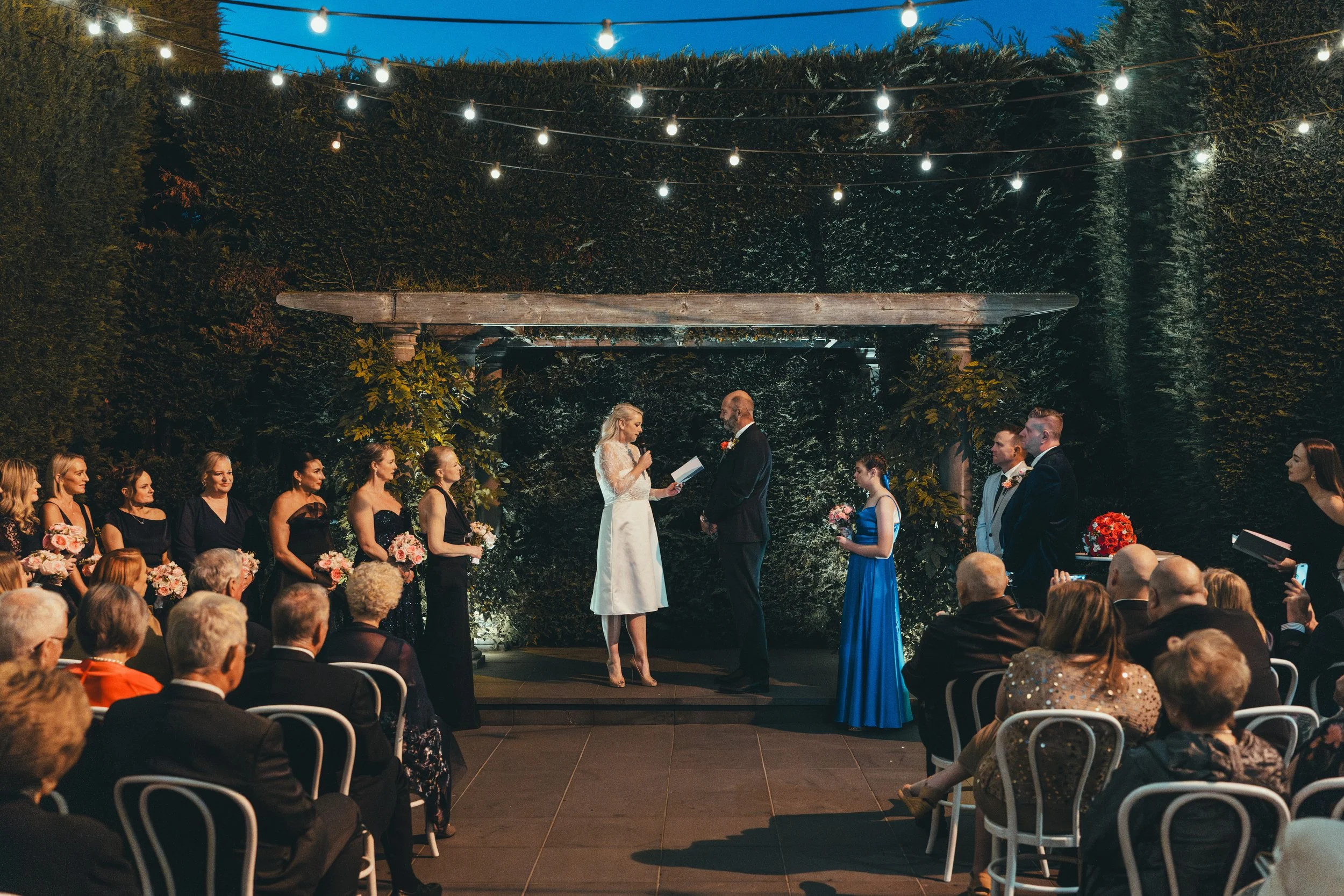 A wedding ceremony taking place outdoors at night with string lights overhead and guests seated watching as the bride and groom exchange vows.