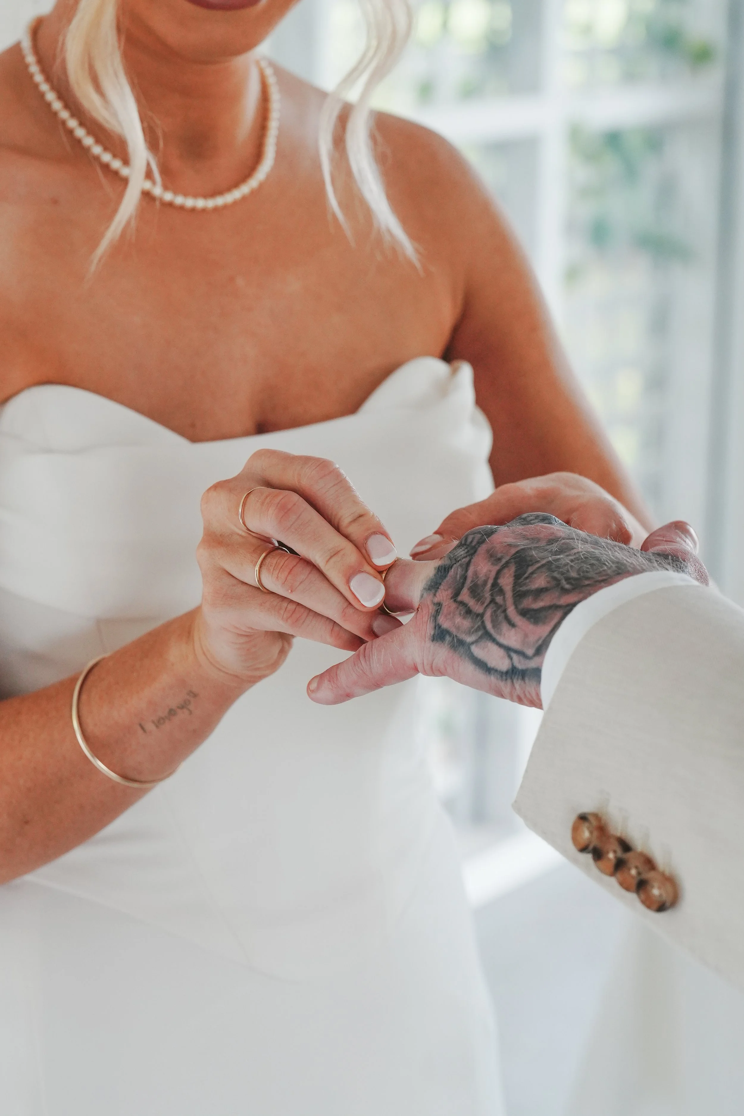 A woman with blonde hair wearing a pearl necklace and white strapless dress is holding hands with a man with a tattooed arm, wearing a white shirt with beads at the sleeve.