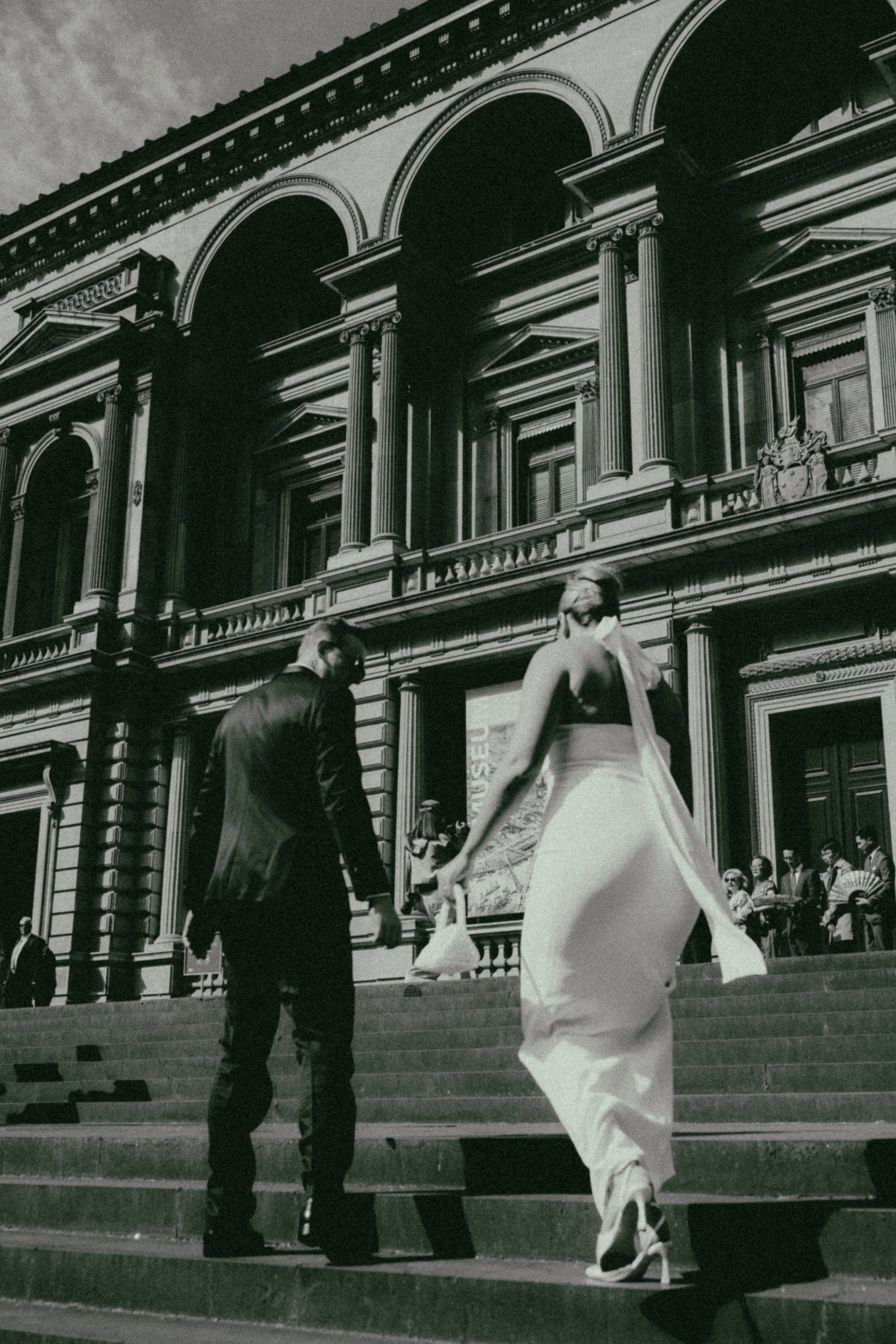 Black and white photo of a woman in a white dress walking up the steps in front of a large ornate building with columns and arched windows, while a man in a suit walks beside her, and other people are gathered in the background.