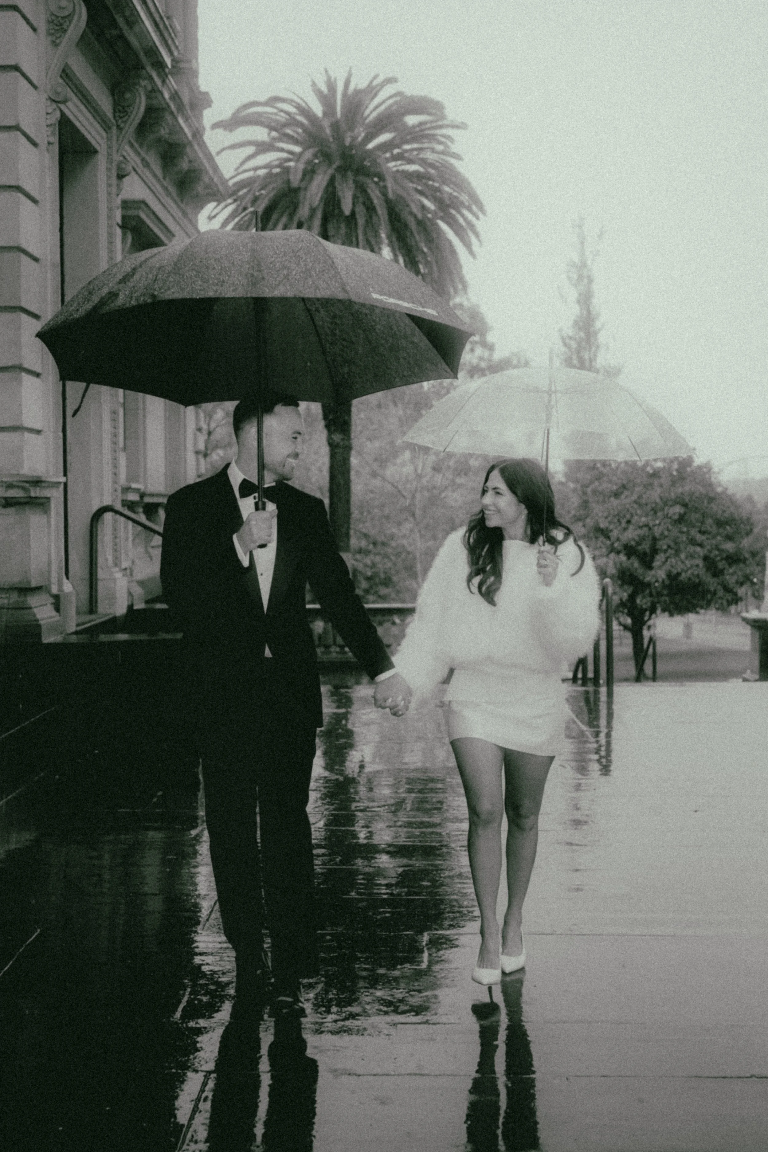 A black and white photo of a man in a tuxedo and a woman in a white dress holding hands and walking outside on a rainy day, with palm trees and a building in the background. They are holding umbrellas and smiling at each other.