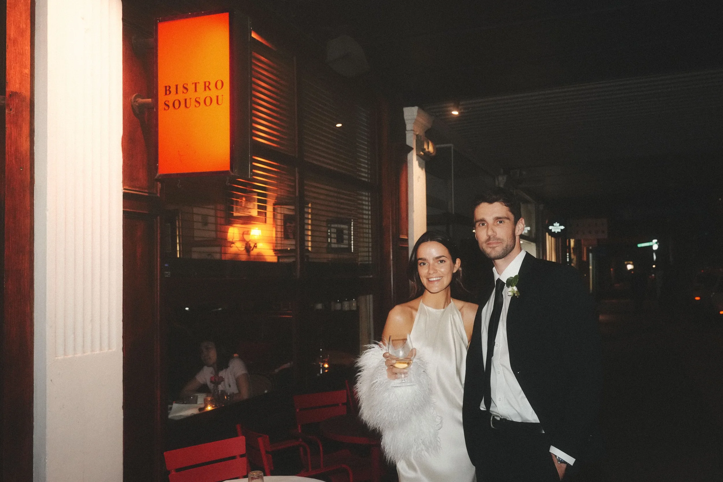A couple dressed in formal attire at a restaurant or bar, with the woman holding a wine glass and the man standing beside her, both smiling.