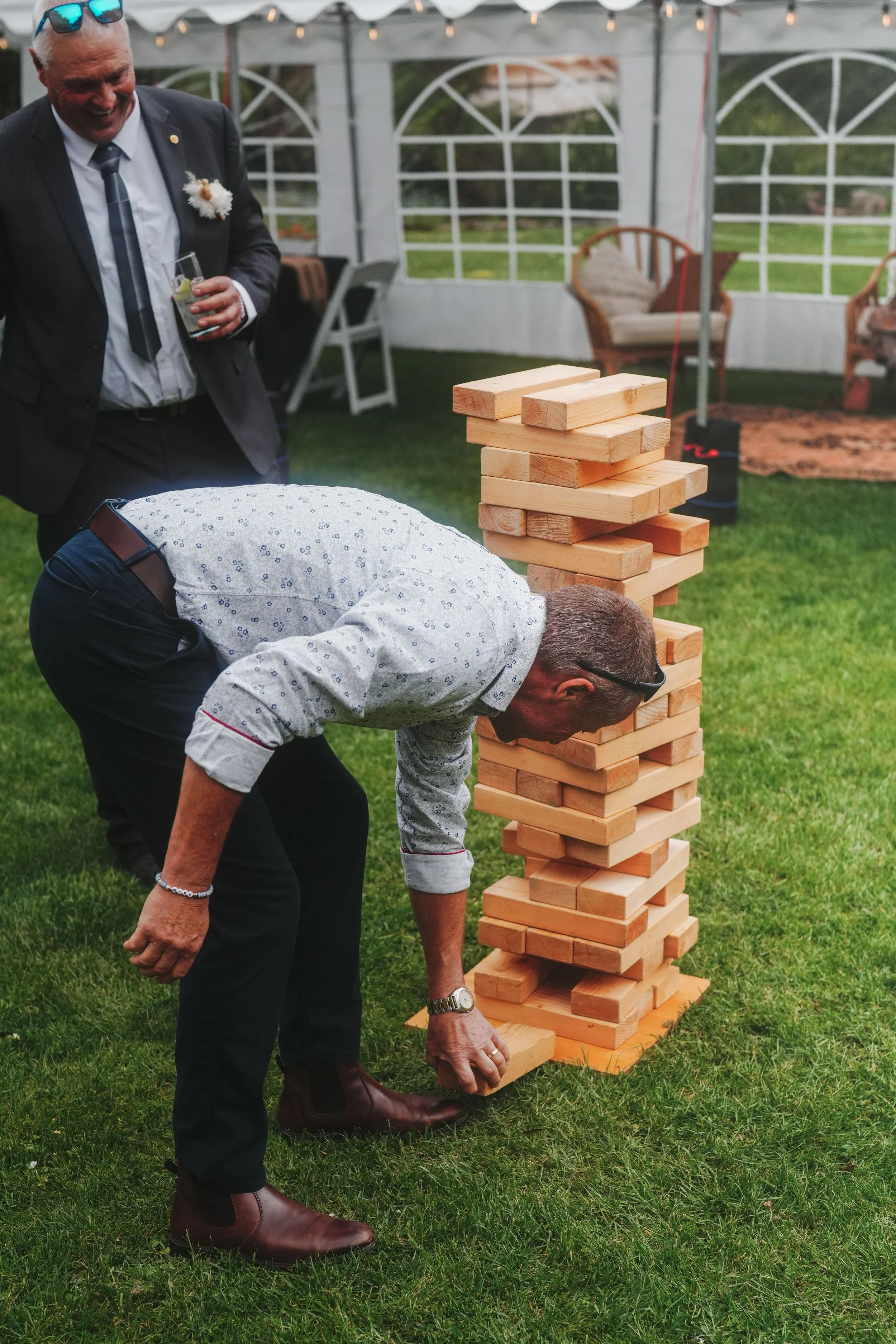 Two men playing large outdoor Jenga game under a tent, with one man bending down and the other smiling, holding a drink, in a backyard setting with chairs and grass.