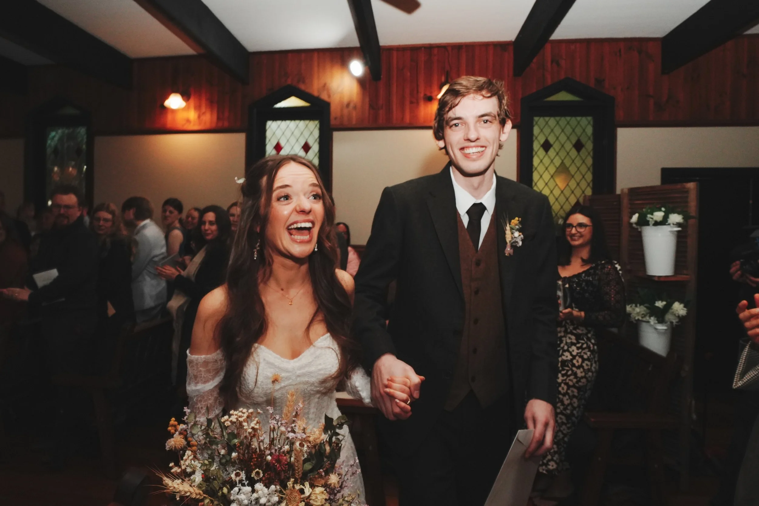 A bride and groom holding hands at their wedding reception, surrounded by guests in a decorated indoor venue.