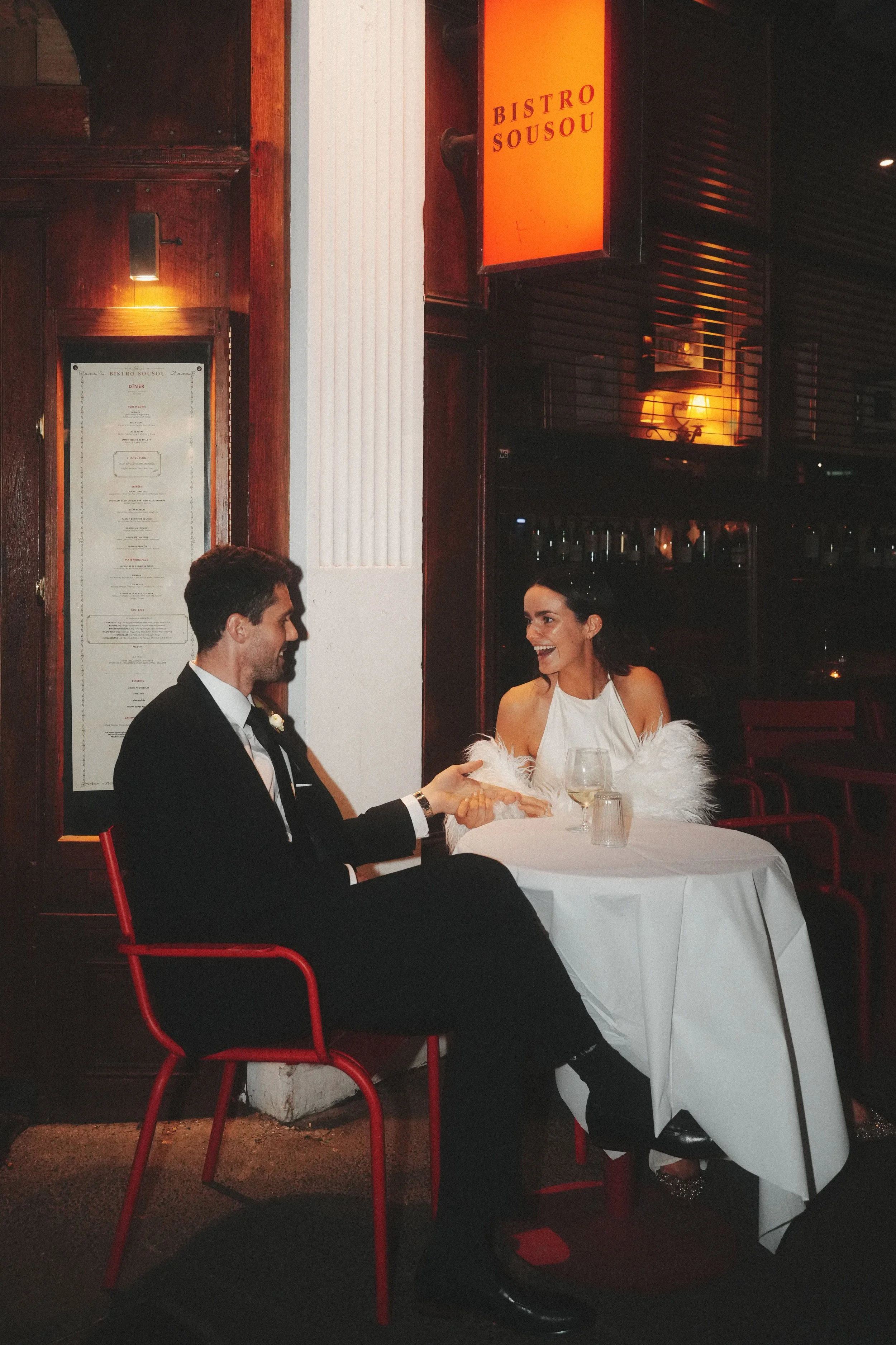 A man in a black suit and a woman in a white dress are sitting at a table in a dimly-lit restaurant named Bistro Sousou. They are smiling and appear to be enjoying a conversation. The table is covered with a white tablecloth and set with glasses and 