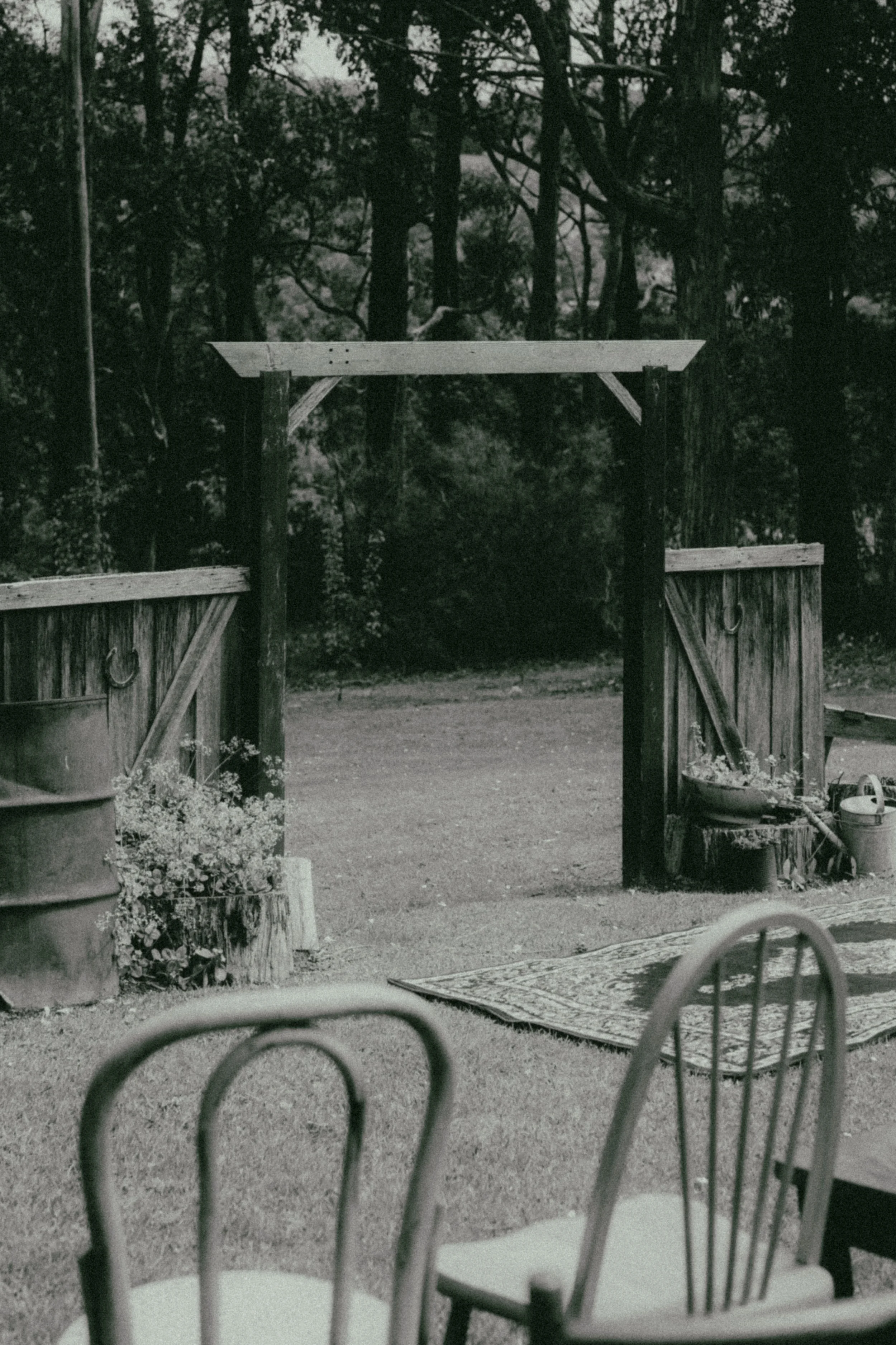A black and white photo of an outdoor scene with trees in the background. In the foreground, there are metal chairs and a table, with some potted plants and garden tools nearby.