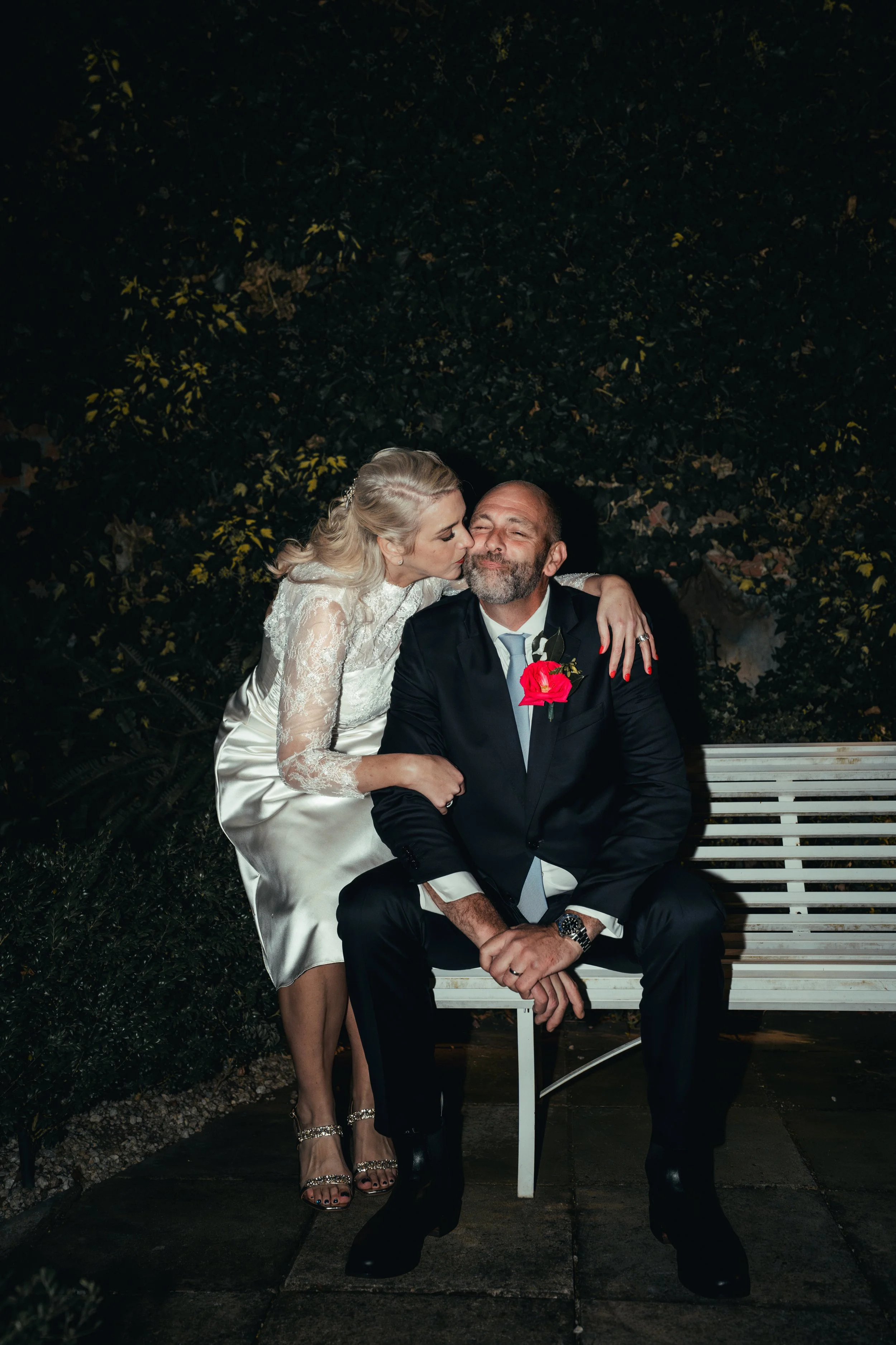 A woman in a lace wedding dress and high-heeled sandals is giving a kiss on the cheek to a smiling man in a dark suit with a boutonniere, sitting on a white bench outside at night, with leafy greenery in the background.