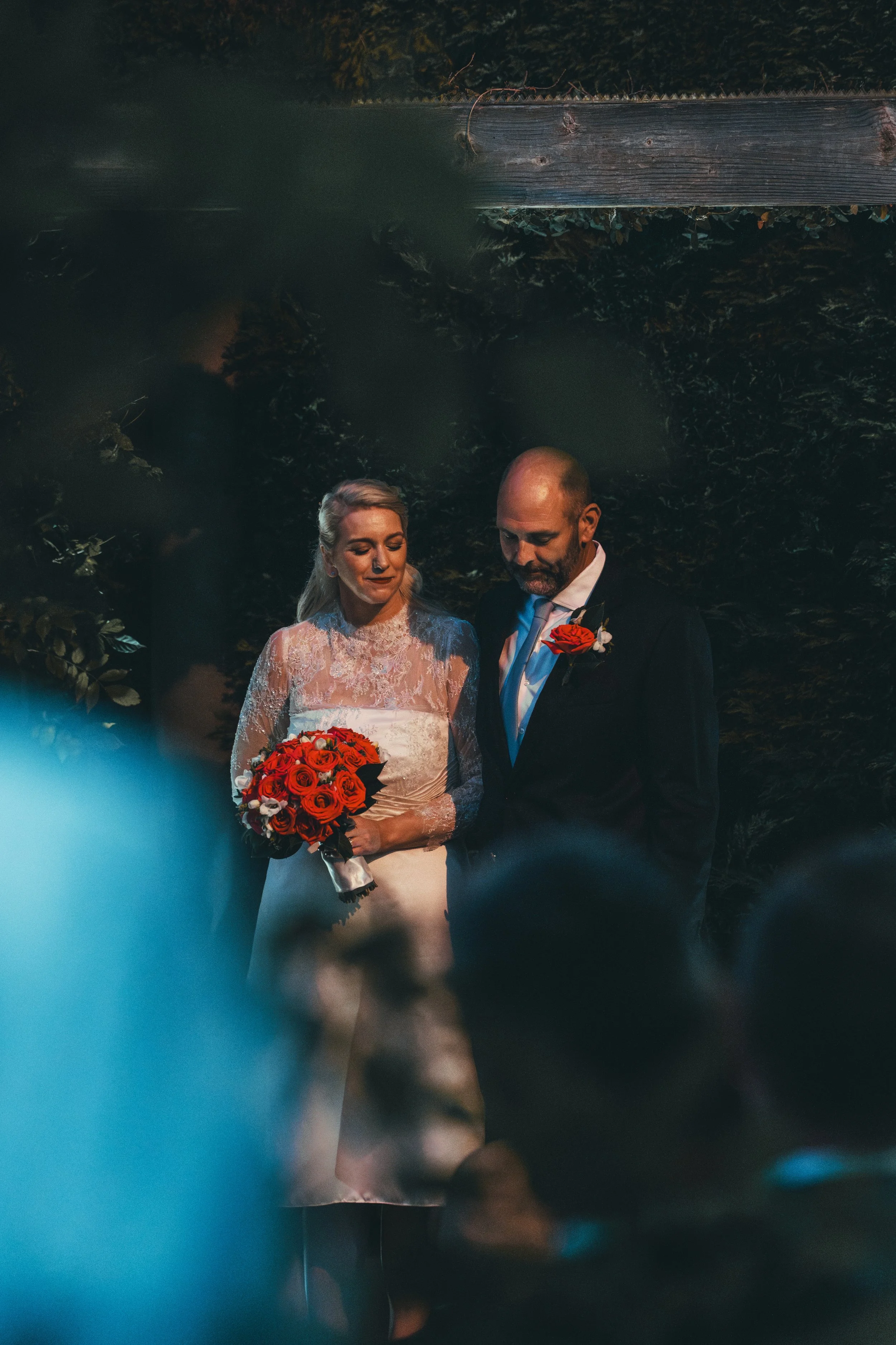 A bride and groom are standing together in front of a dark, leafy background. The bride is holding a bouquet of red roses and is wearing a white lace wedding dress with long sleeves. The groom is dressed in a dark suit with a white shirt and a bouton