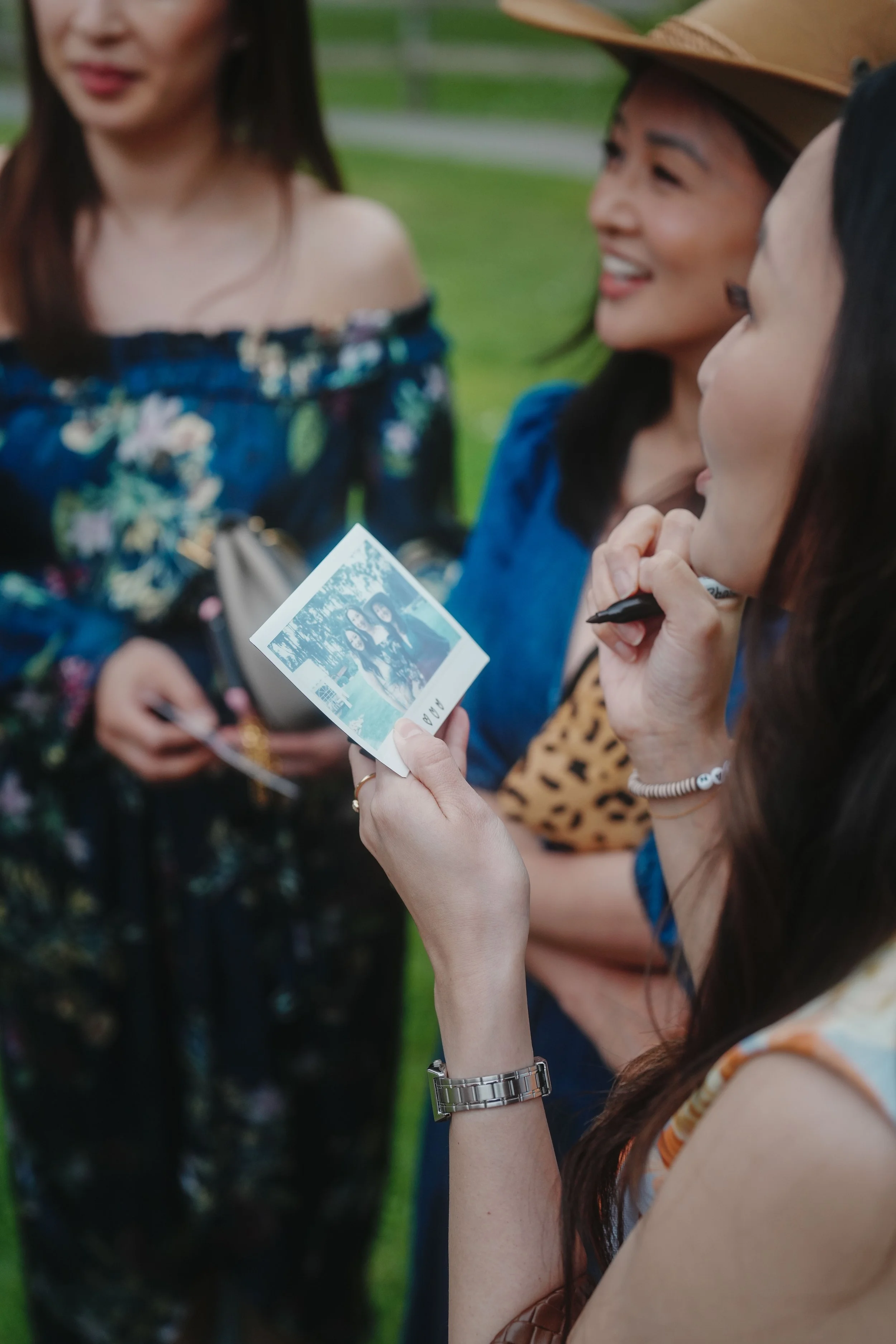 Three women outdoors, one holding a printed photo, smiling and engaging in conversation. The woman in the foreground has long brown hair, wears a sleeveless top, a silver watch, and bracelets. Two women in the background are smiling, one wearing a wi