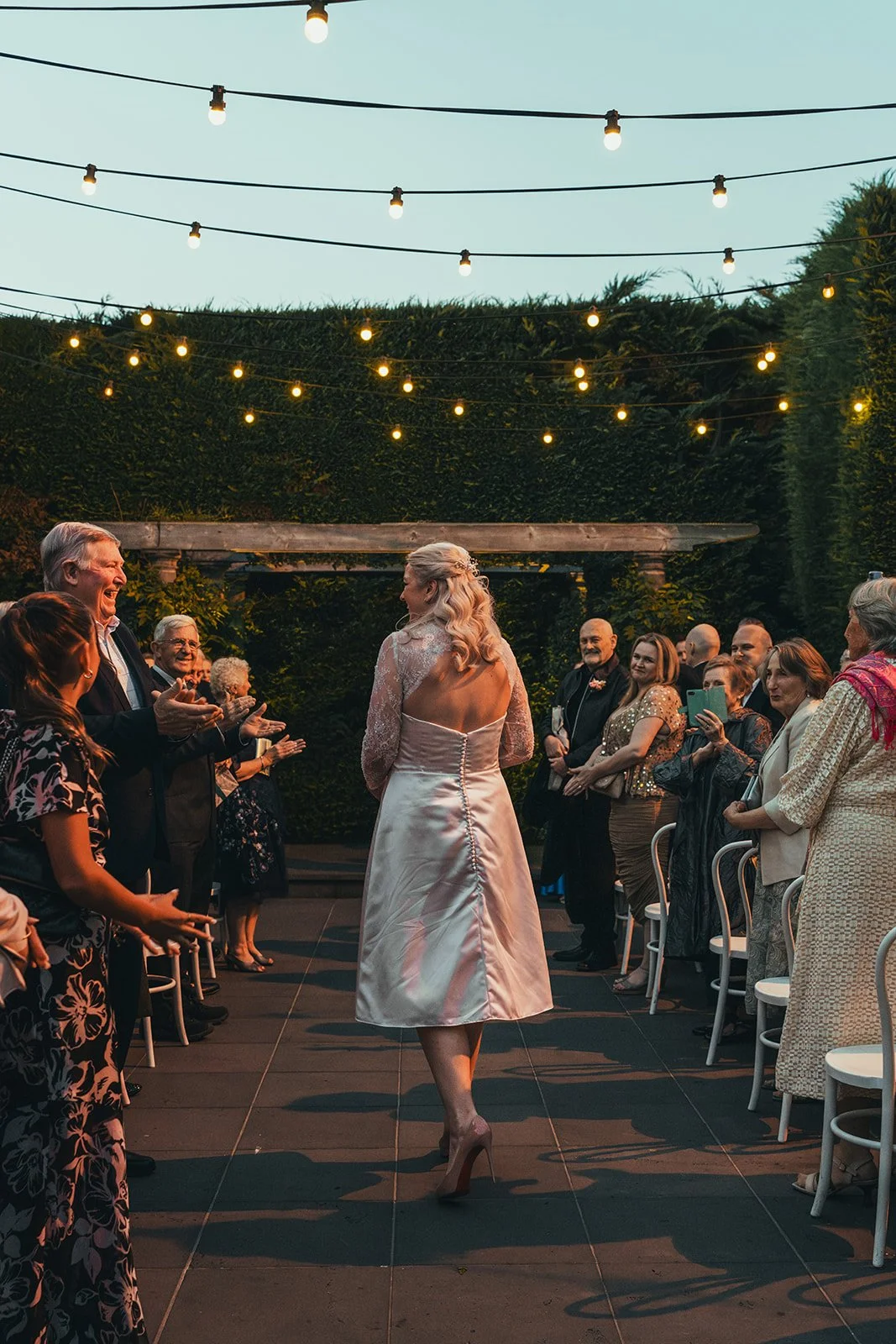 Bride walking down the aisle at an outdoor wedding reception during the evening, surrounded by guests under string lights.