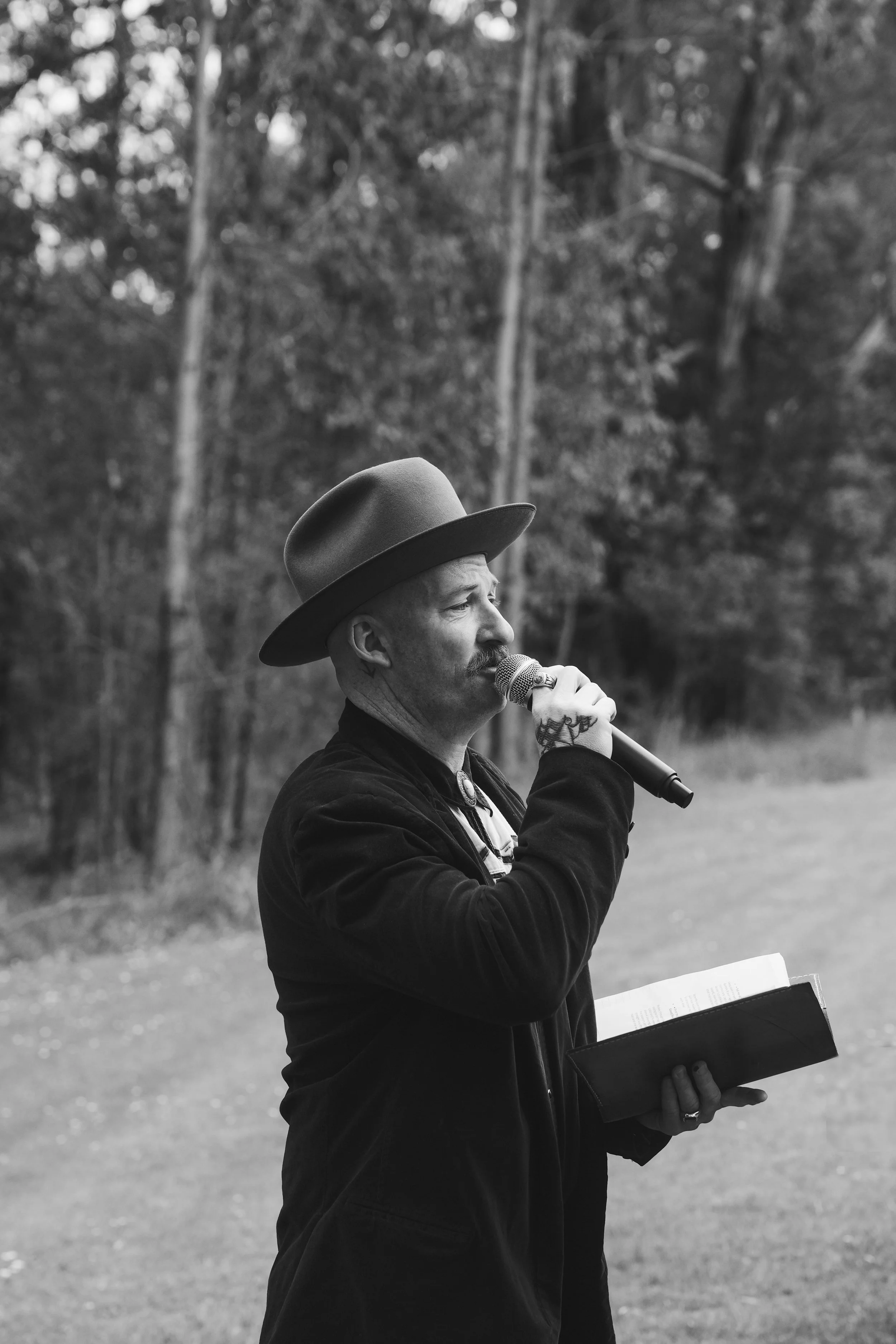 A man wearing a hat and black clothing is standing outdoors in a forest, singing into a microphone while holding a music sheet or book.