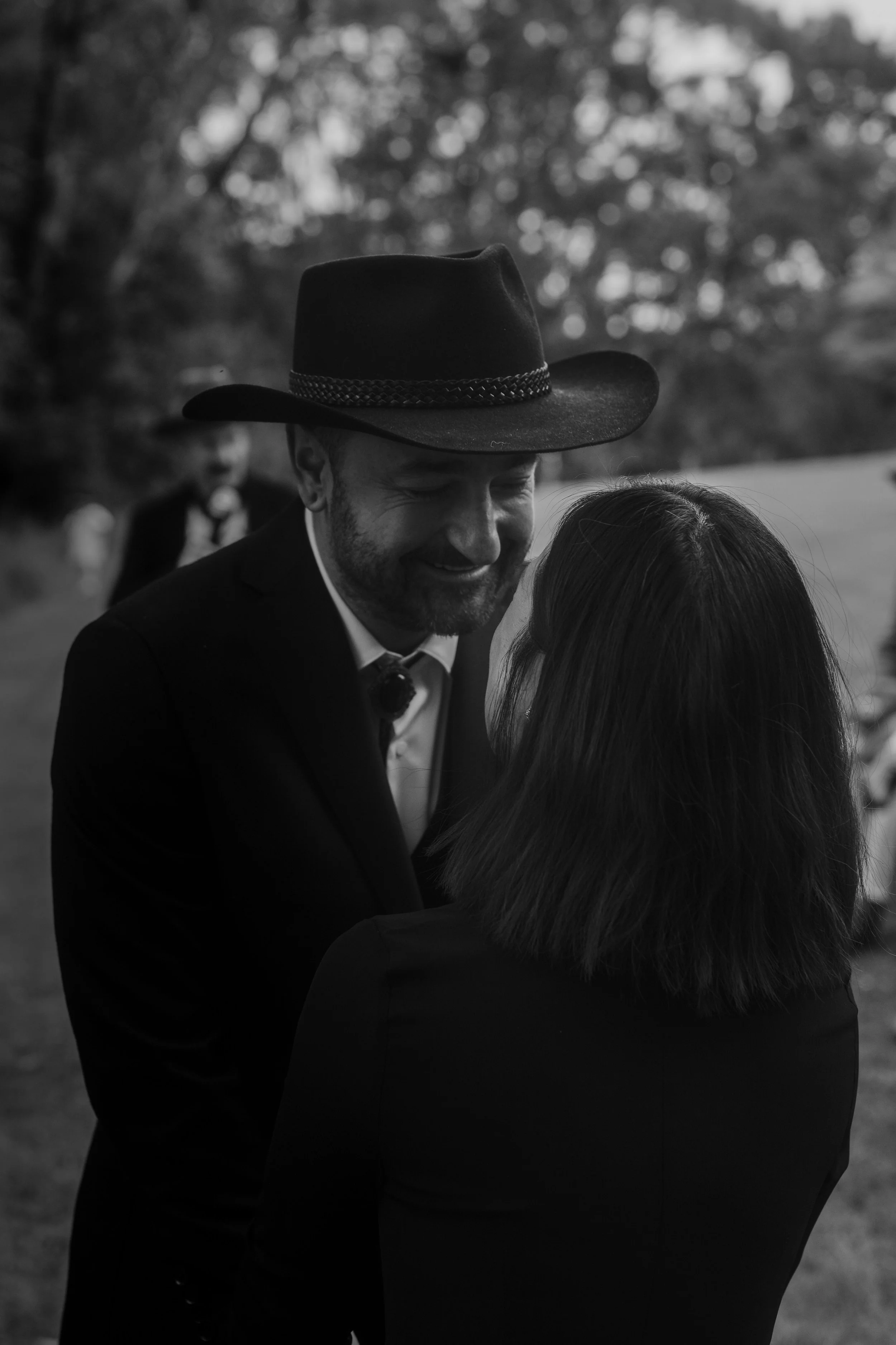A black and white photo of a couple embracing outdoors, with the man wearing a cowboy hat and the woman with long hair, smiling and leaning into each other.
