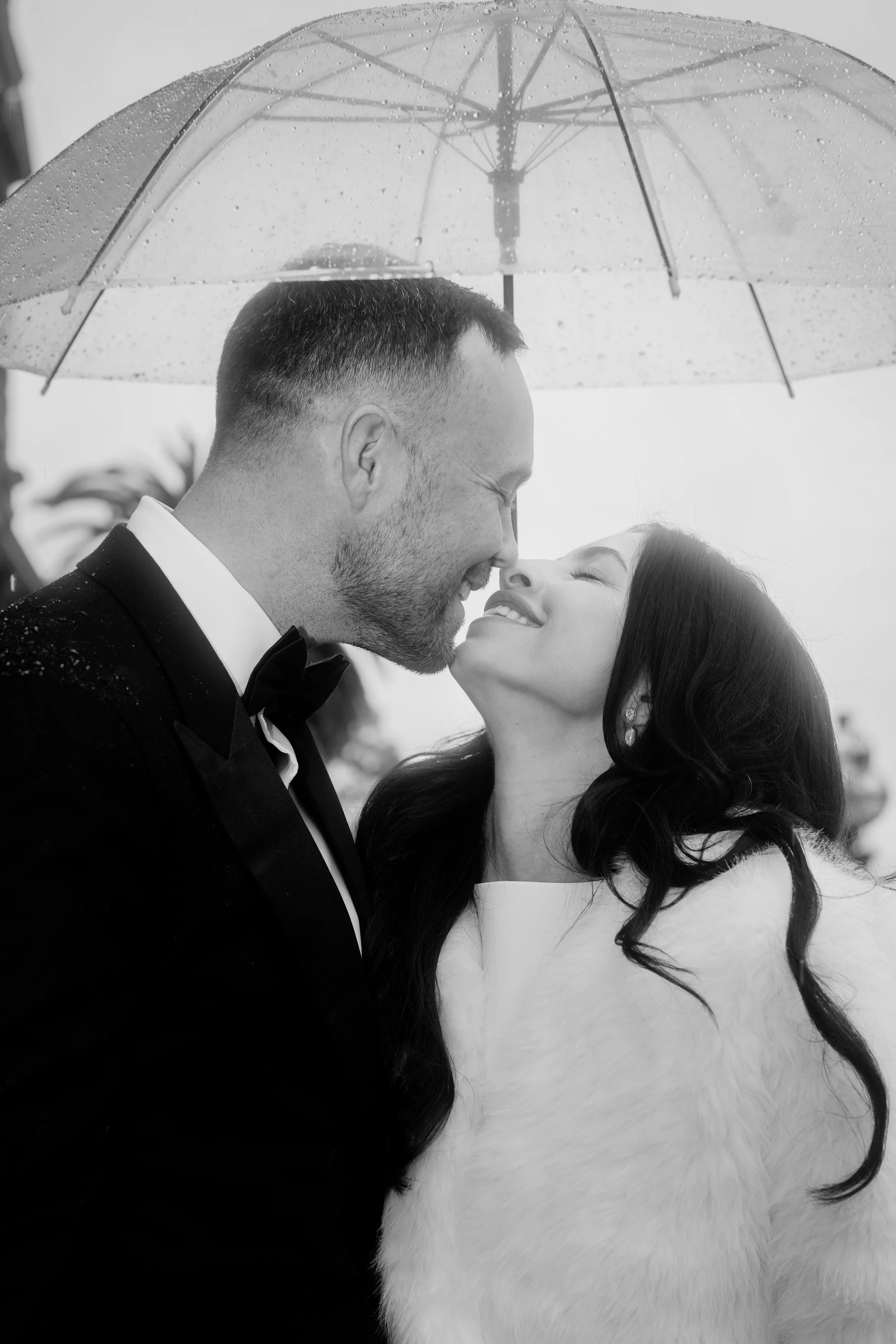 A black-and-white photo of a couple under a transparent umbrella, smiling at each other. The man is dressed in a tuxedo and the woman is wearing a white dress with long, dark hair. Raindrops are visible on the umbrella.