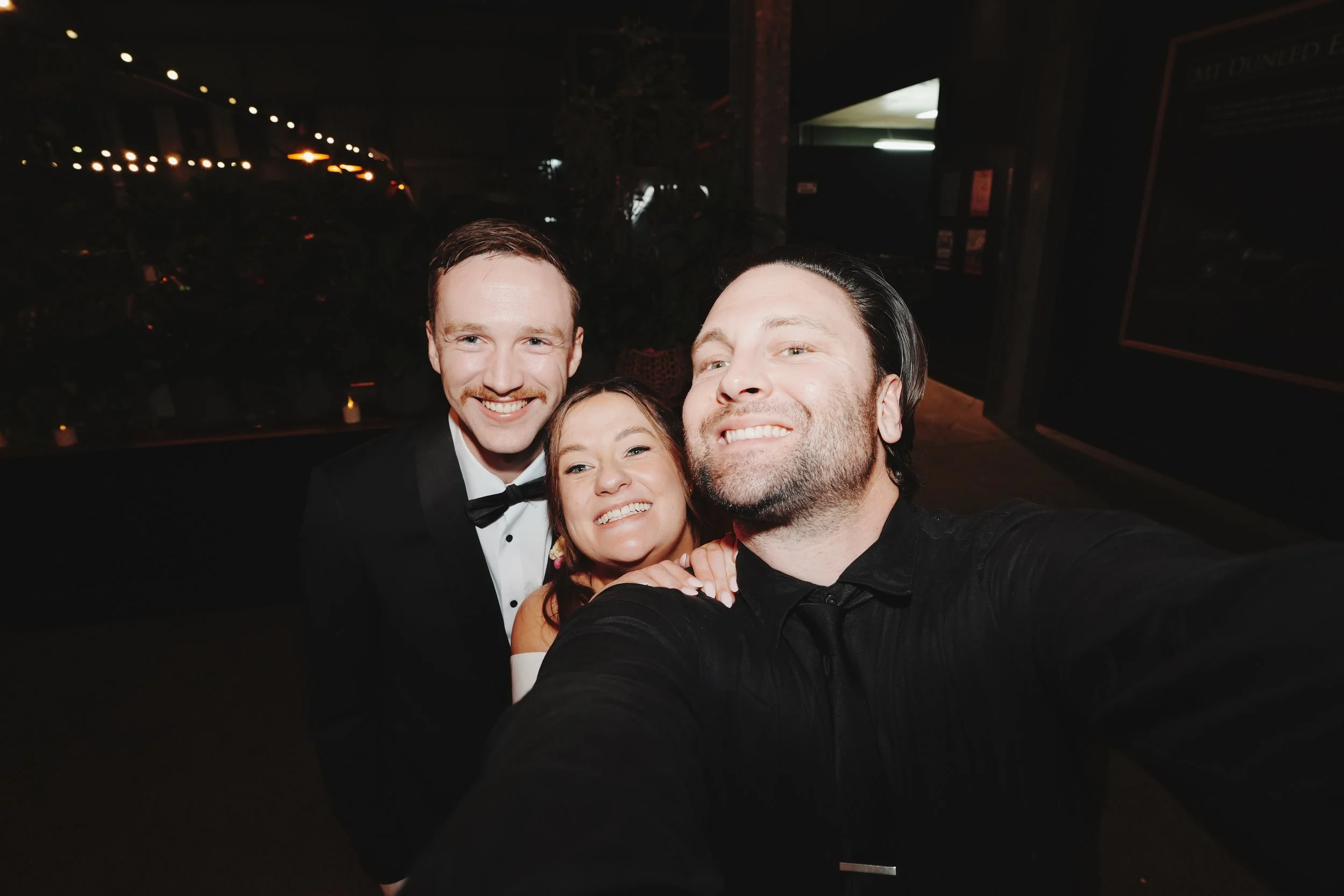 Three friends smiling and taking a selfie together at night, dressed formally with two men in tuxedos and a woman in a white dress. Behind them are dark surroundings and some lights.