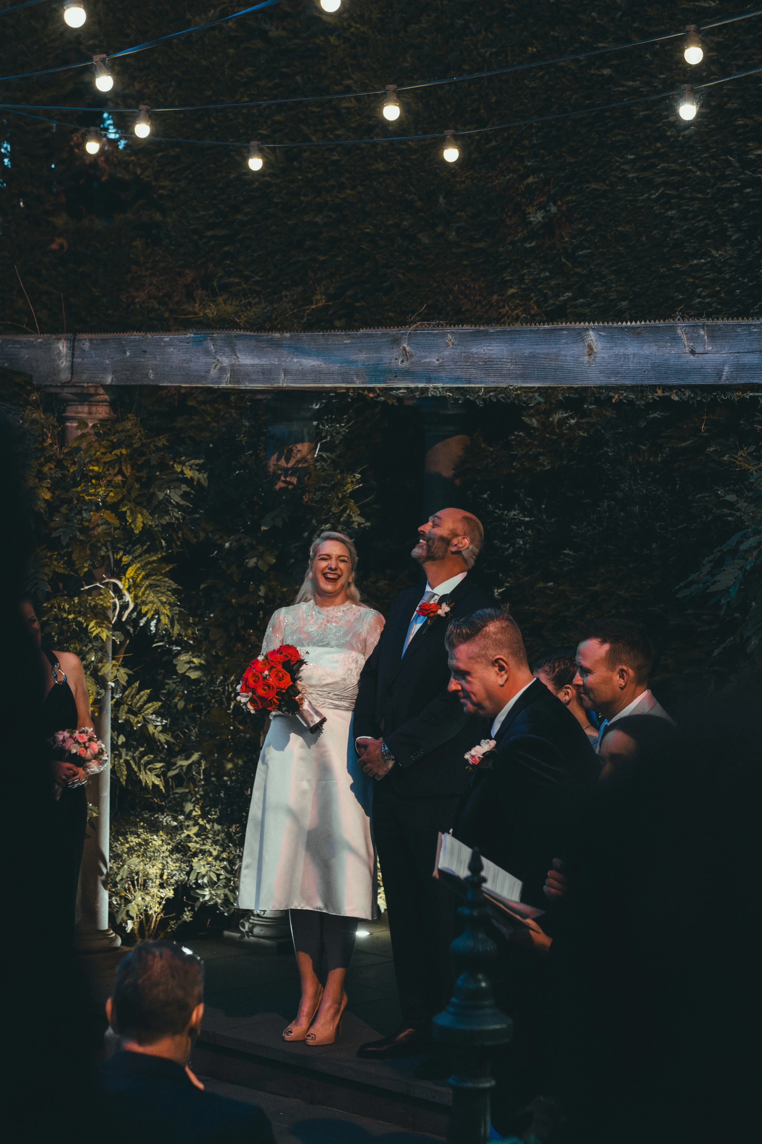 A wedding ceremony taking place outdoors at night under string lights. The bride is holding a bouquet of red roses and smiling, standing next to the groom who is dressed in a dark suit. Guests are seated around, some holding program sheets, and are w