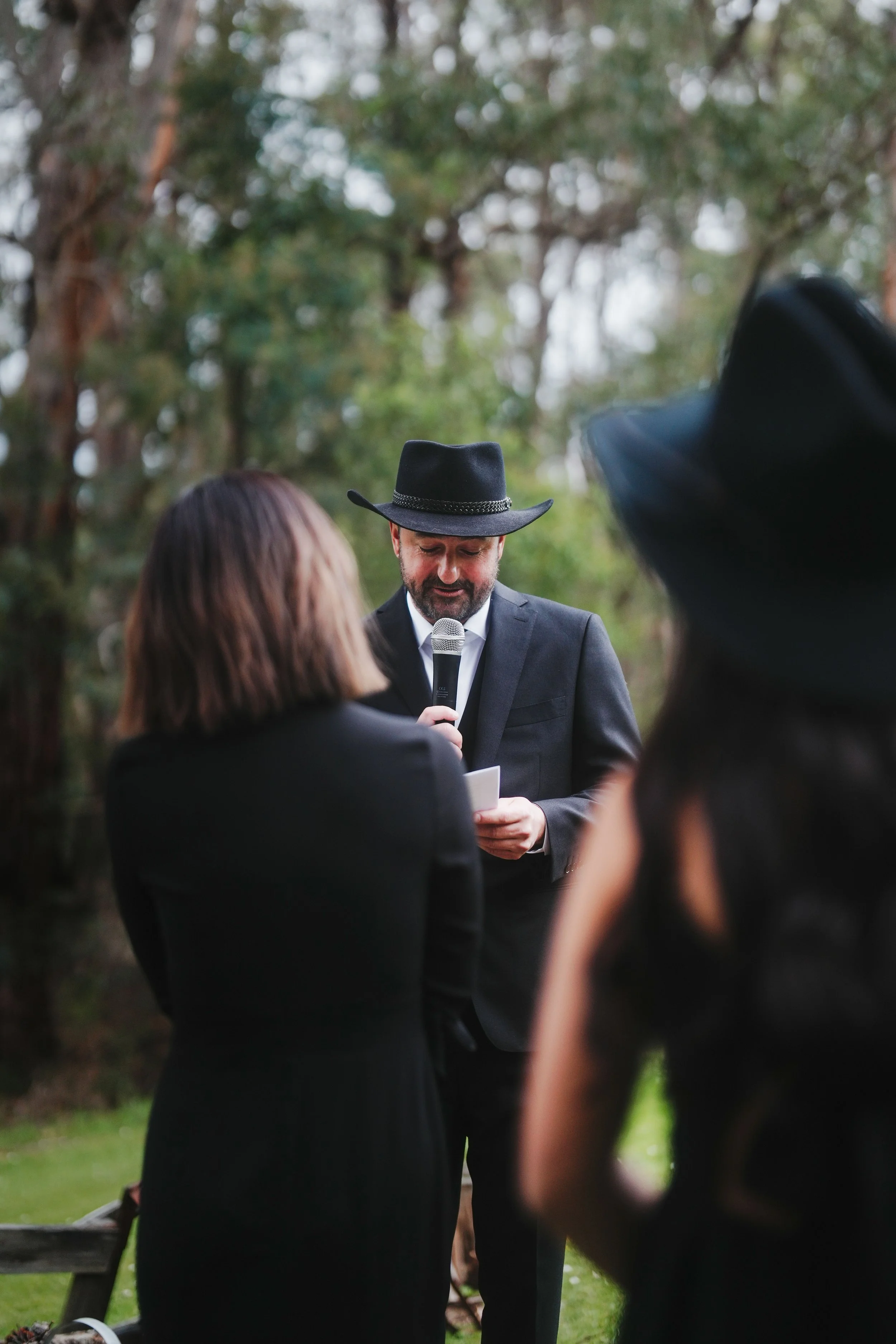 A man reading a speech at an outdoor event, wearing a black cowboy hat and suit, surrounded by people, with trees in the background.