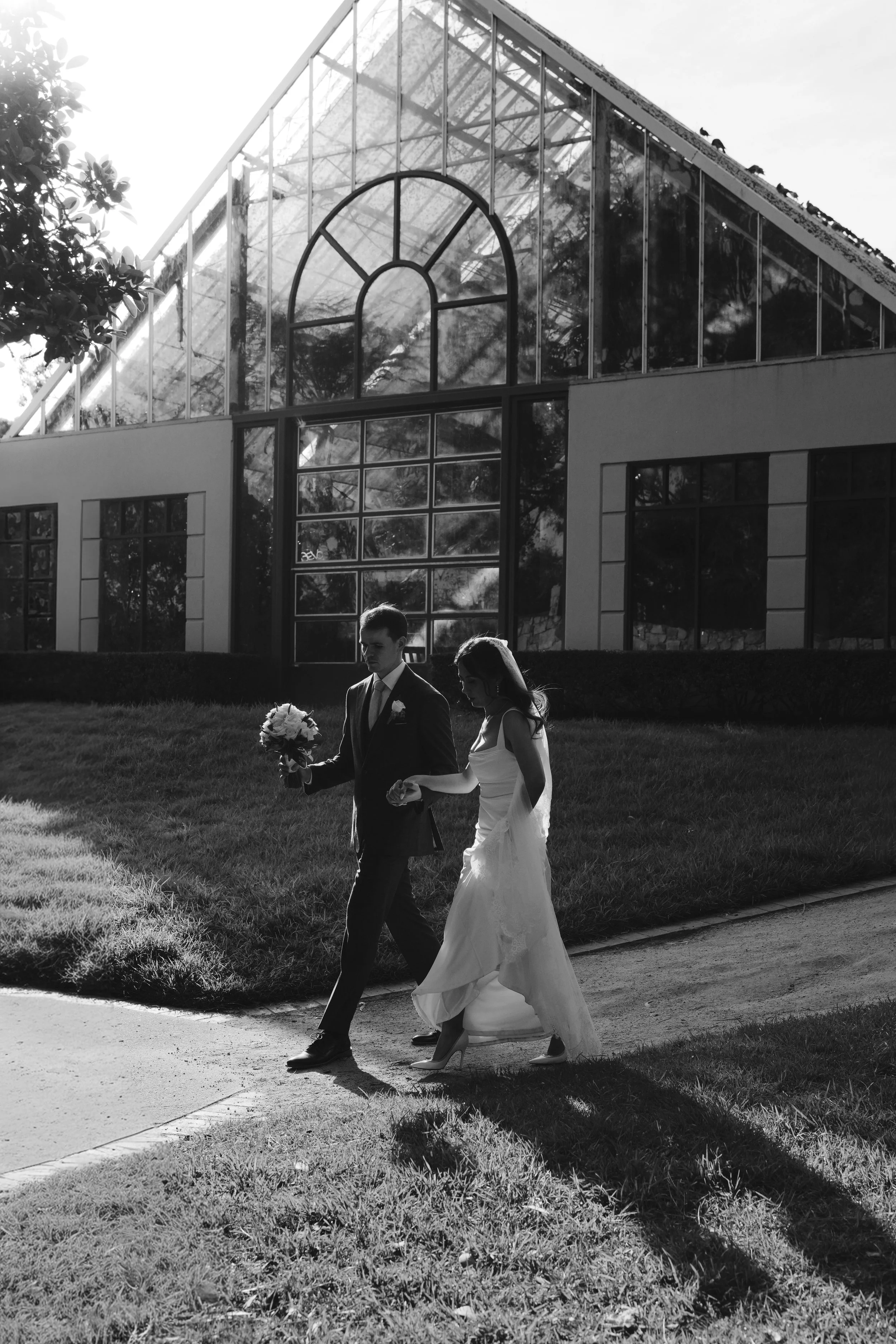 A black and white photo of a bride and groom walking outside on a sidewalk in front of a modern glass building, with the bride wearing a long wedding dress and holding a bouquet, and the groom dressed in a suit with a boutonniere.