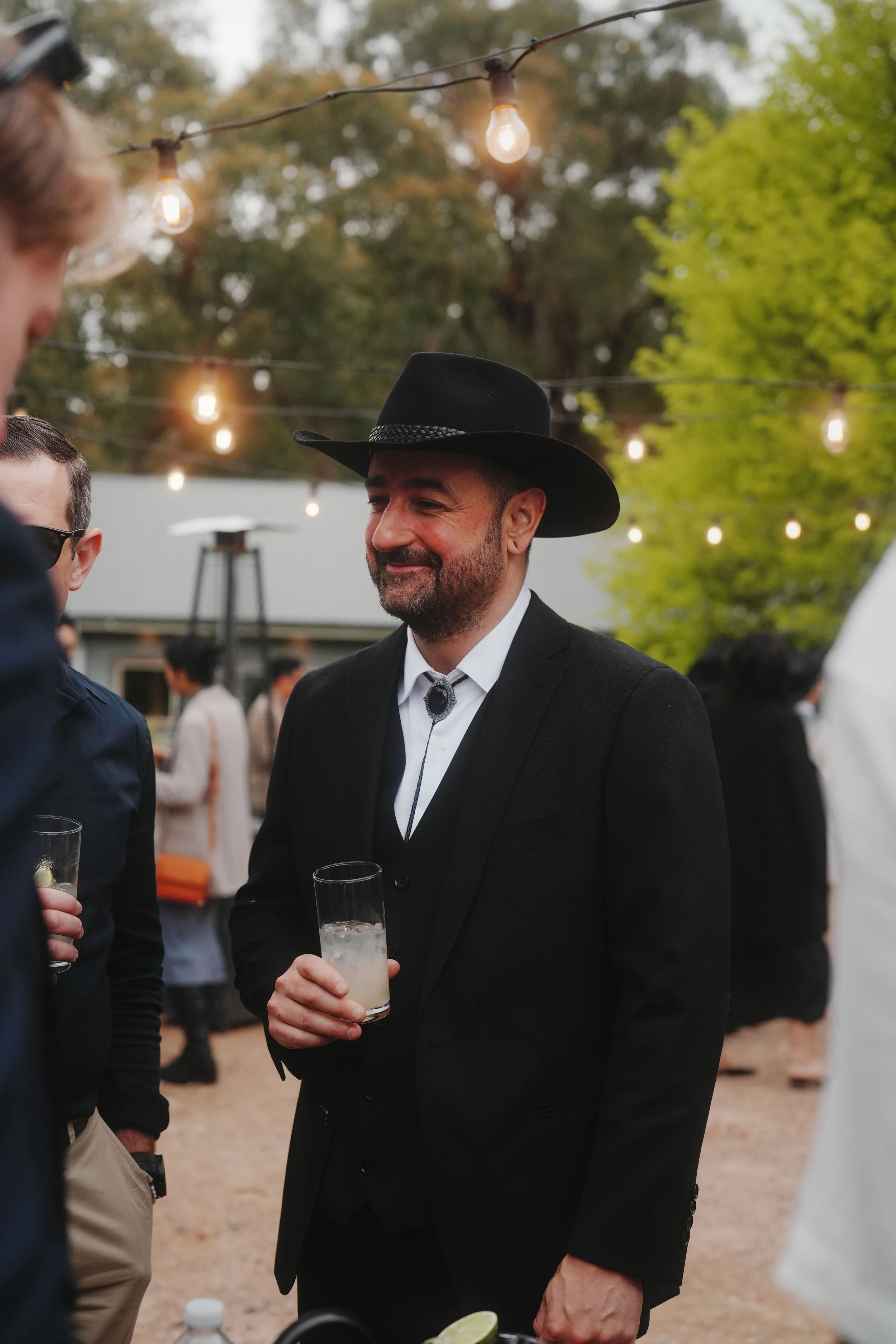 Man with a beard and mustache wearing a black cowboy hat and a black suit jacket, holding a drink with ice and a lime wedge, at an outdoor gathering with string lights and people in the background.