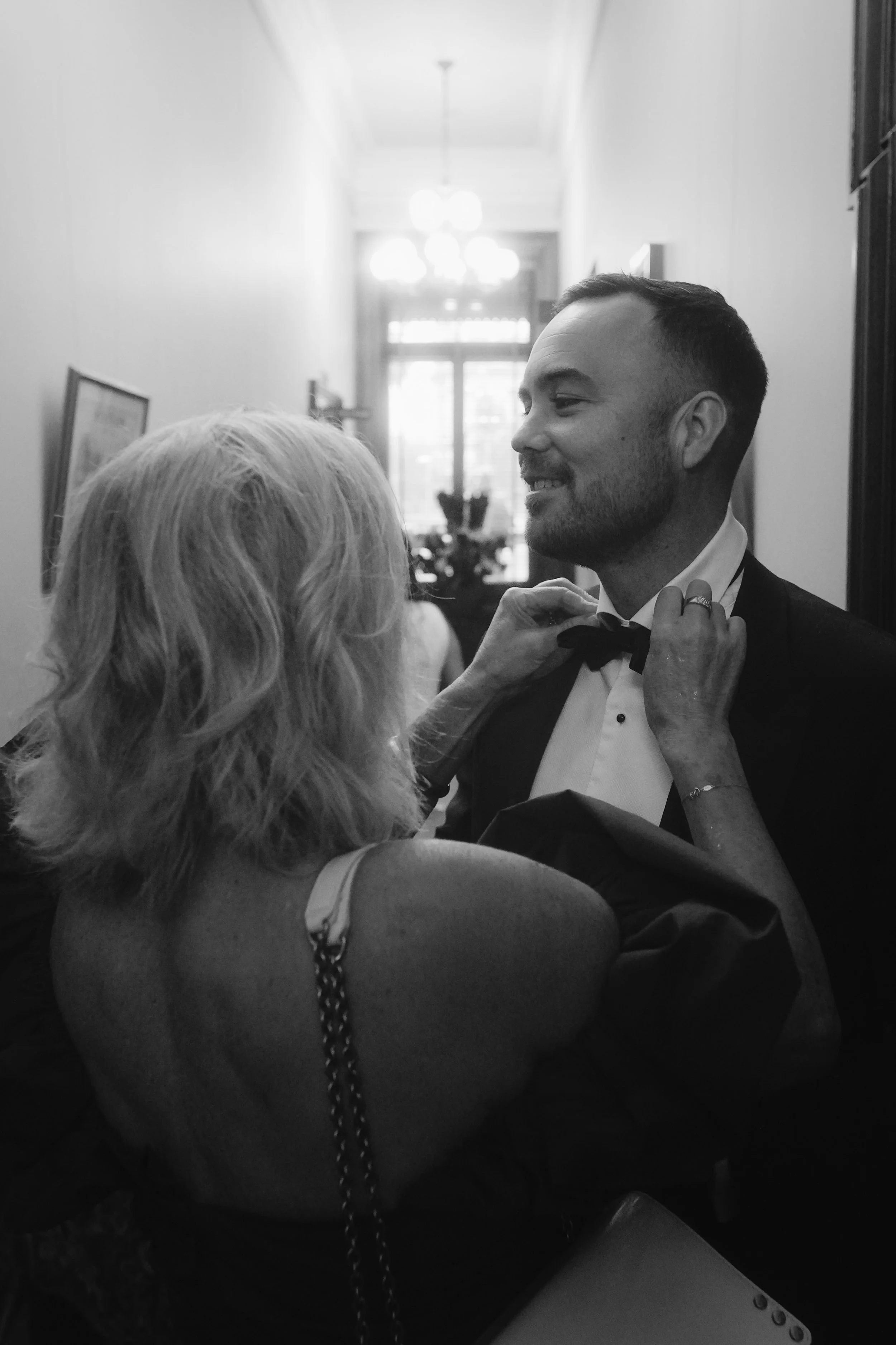 A black and white photo of a man in a tuxedo smiling as an elderly woman adjusts his bow tie in a hallway with hanging lights and framed pictures on the wall.
