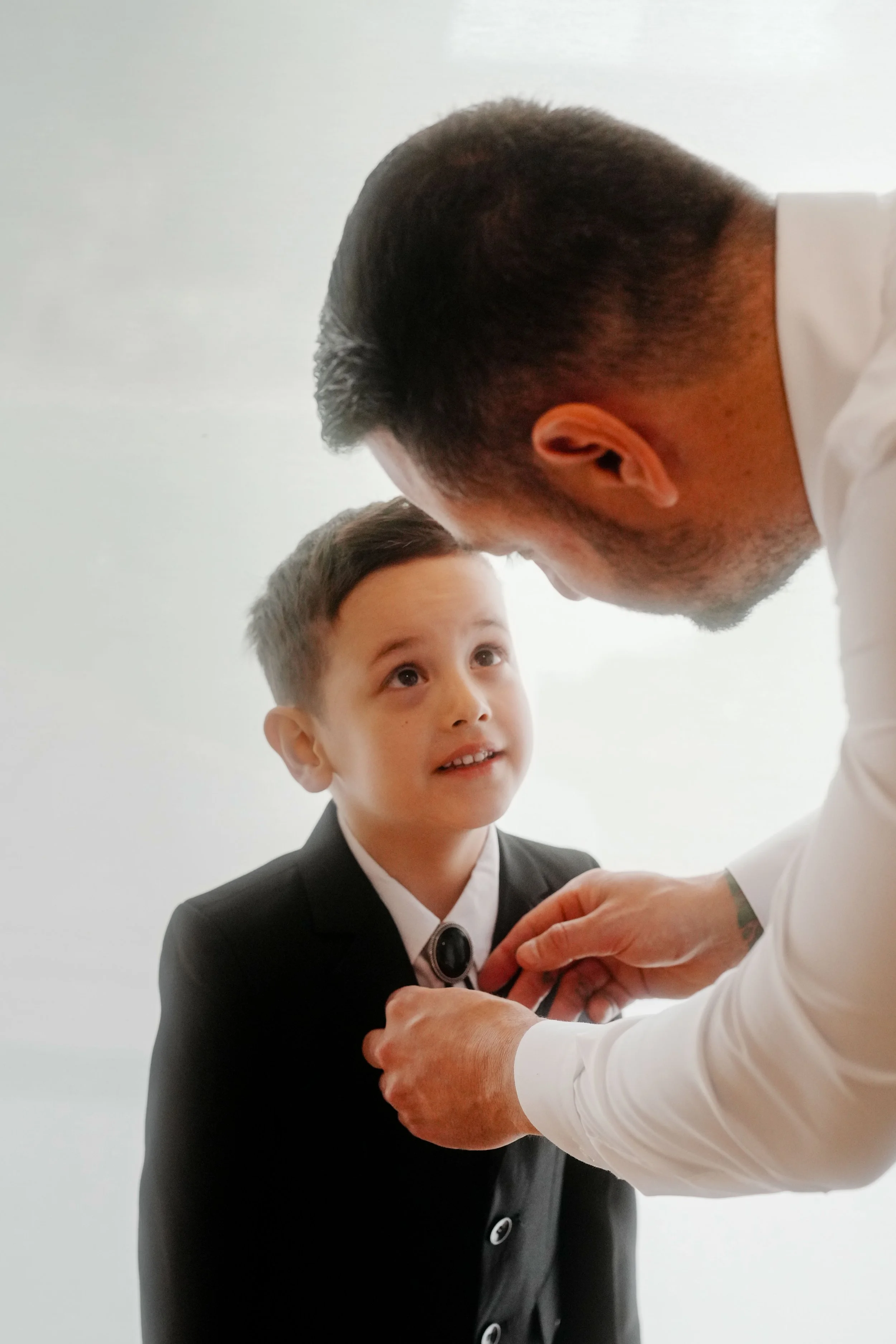 A young boy in a formal black suit and white shirt looking up at an adult man, possibly a doctor or official, who is placing a pin or medal on his shirt during a ceremony or event.