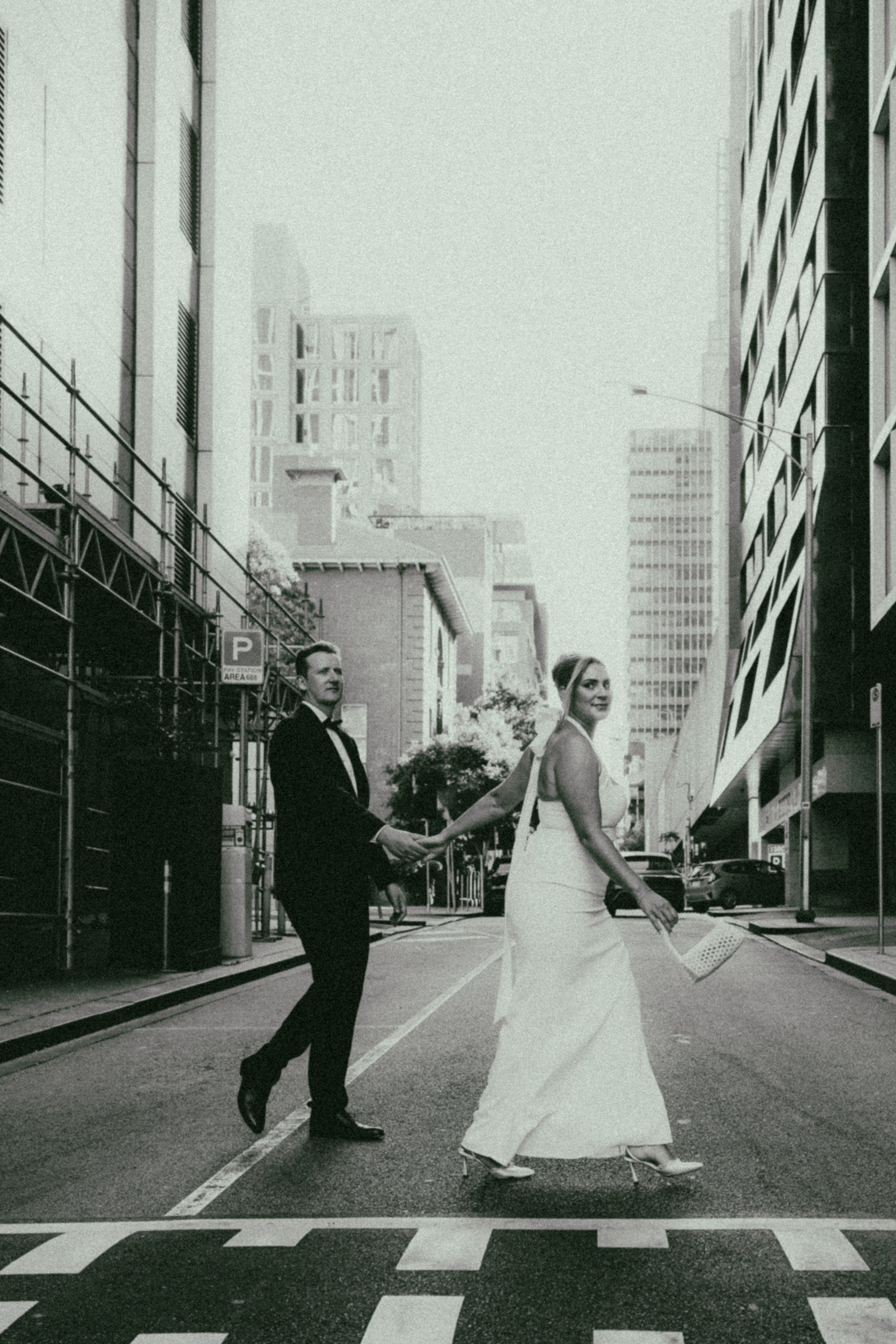 A wedding couple crossing a city street on their wedding day, holding hands and smiling. The groom is in a black tuxedo, and the bride is in a white gown with high heels, holding a bouquet of flowers and a paper fan.