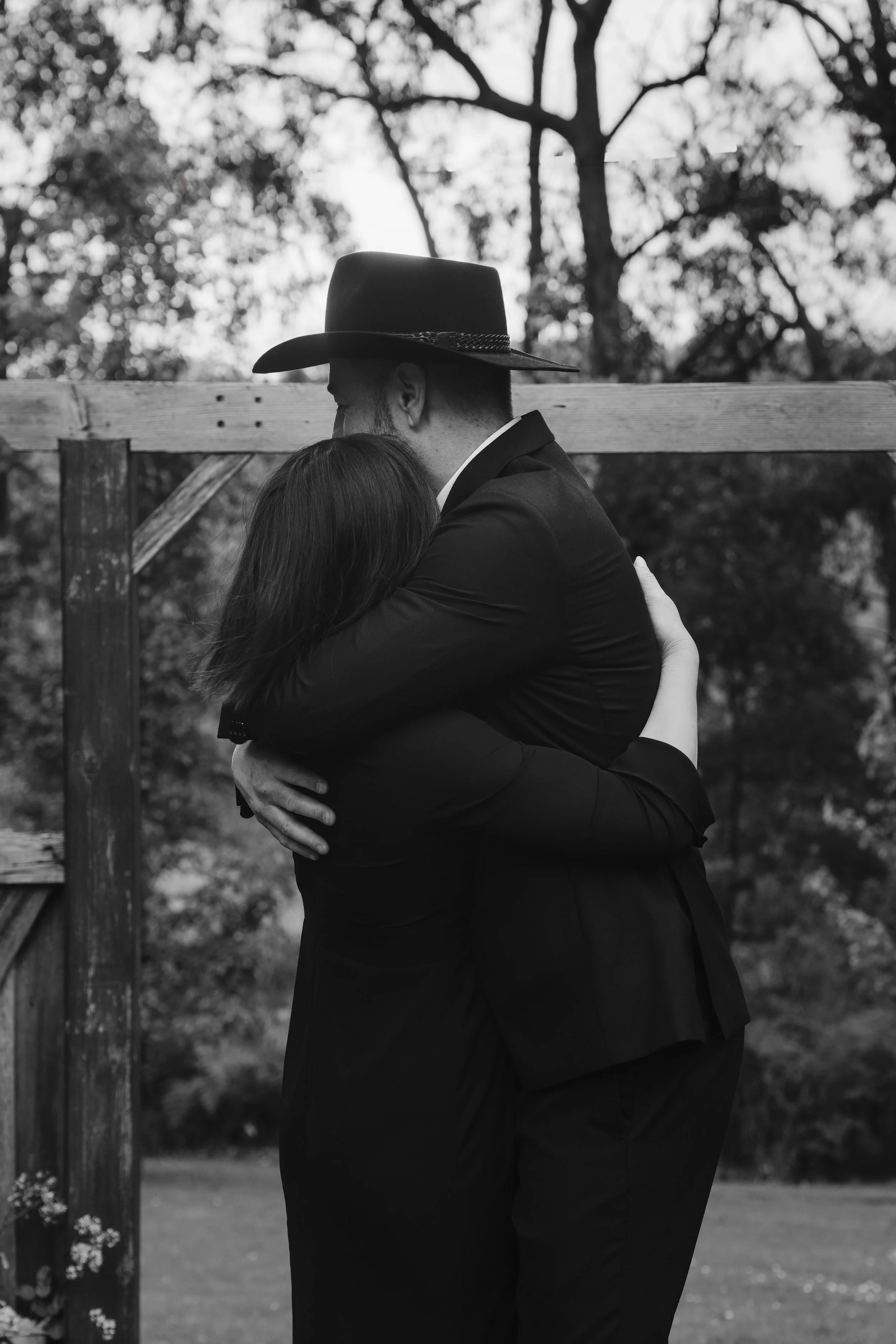 Black and white photo of a couple embracing outdoors, the woman with long hair and the man wearing a hat, with trees in the background.