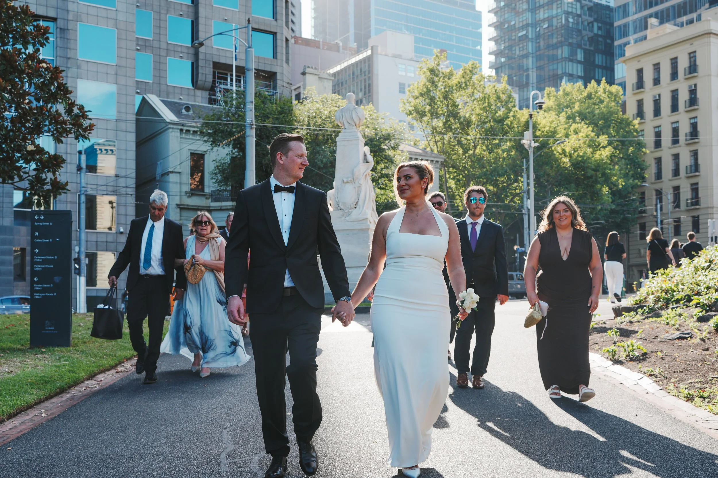 A wedding couple walking hand in hand with guests behind them in an urban park setting with tall buildings and trees.
