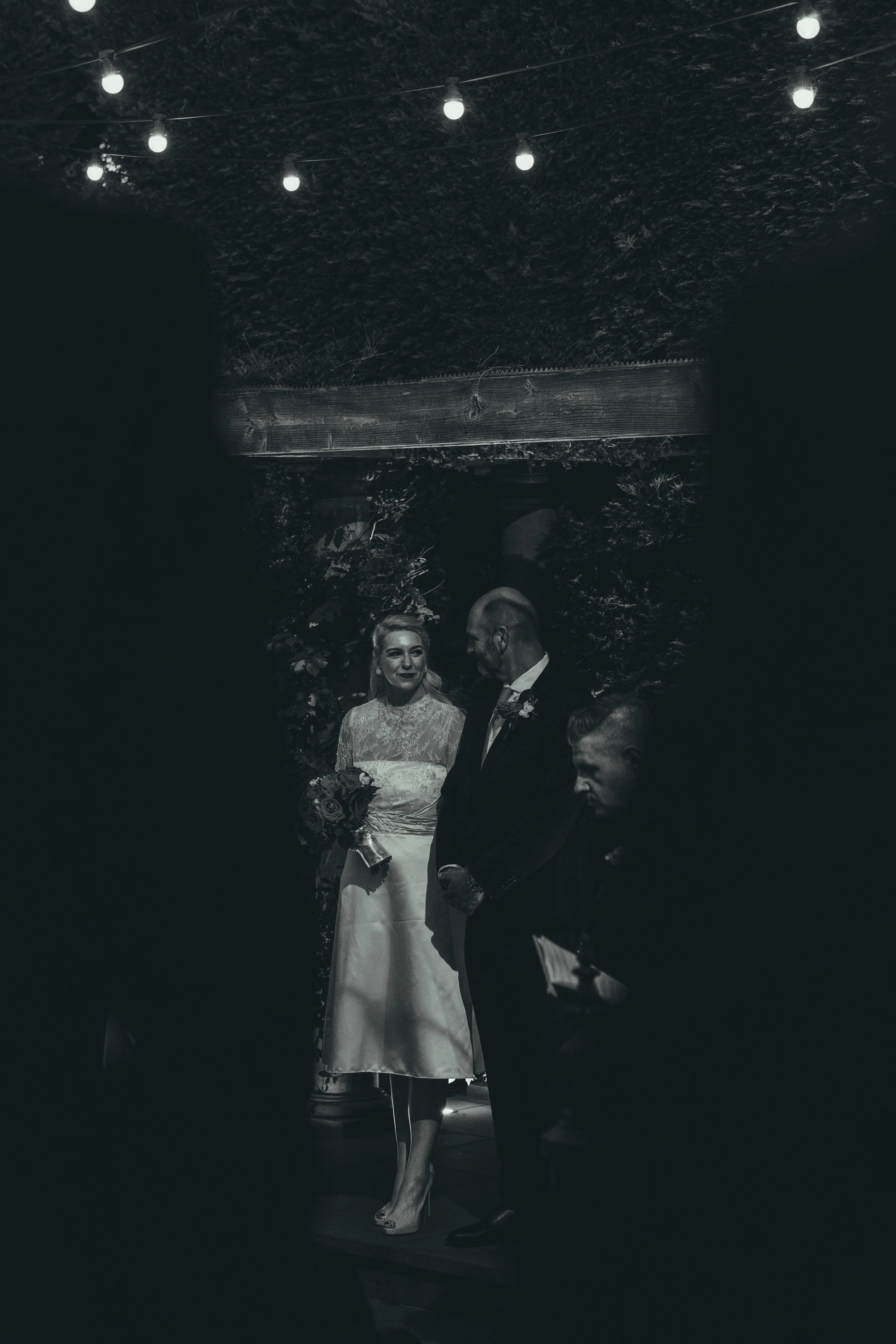 A bride and groom standing on a stage during their wedding ceremony, surrounded by darkness with hanging string lights overhead.