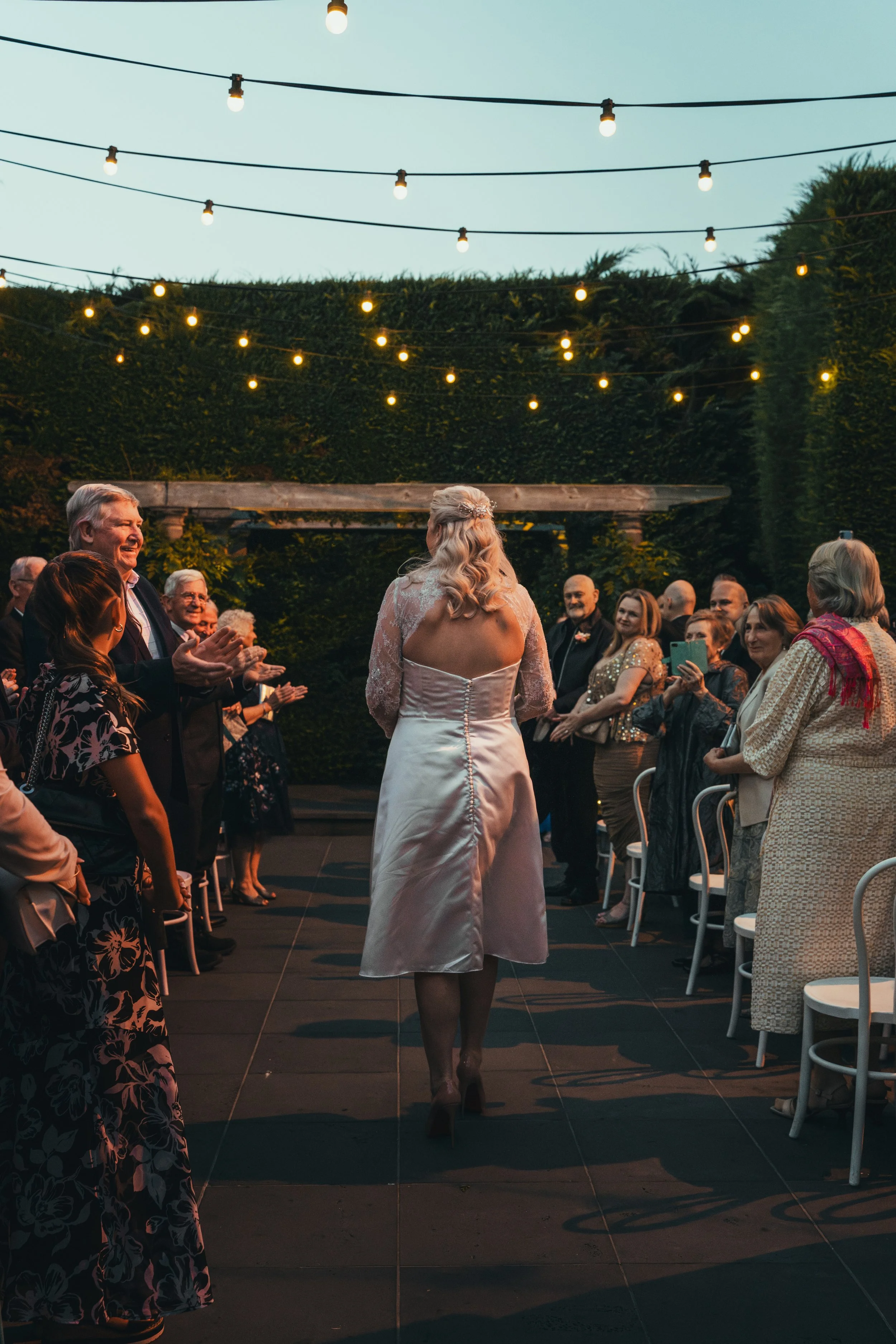 A wedding ceremony taking place outdoors at dusk, with a bride walking down a path lined with seated guests, string lights overhead, and lush greenery background.