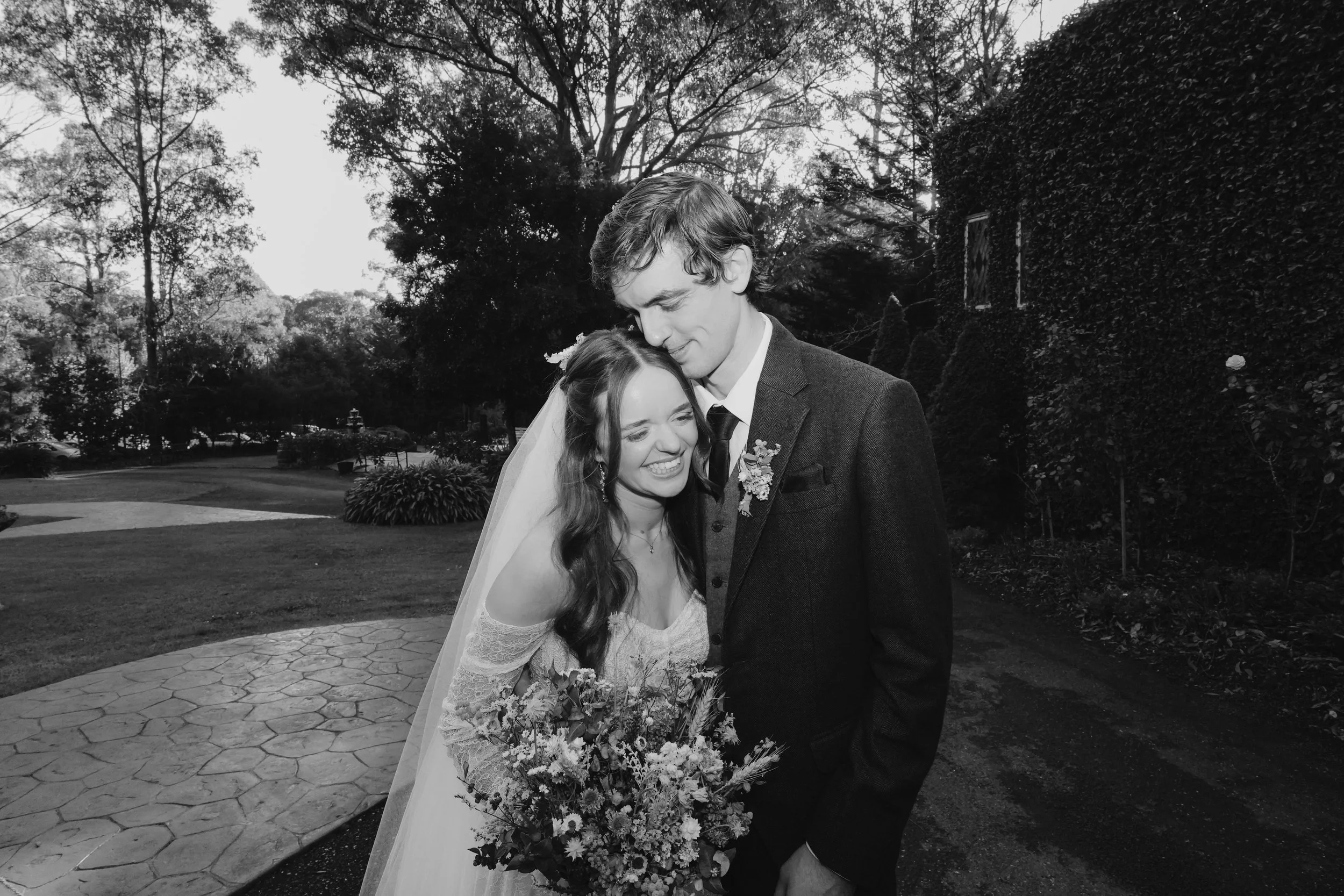 A black and white photo of a bride and groom embracing outdoors, smiling, with trees and bushes in the background.