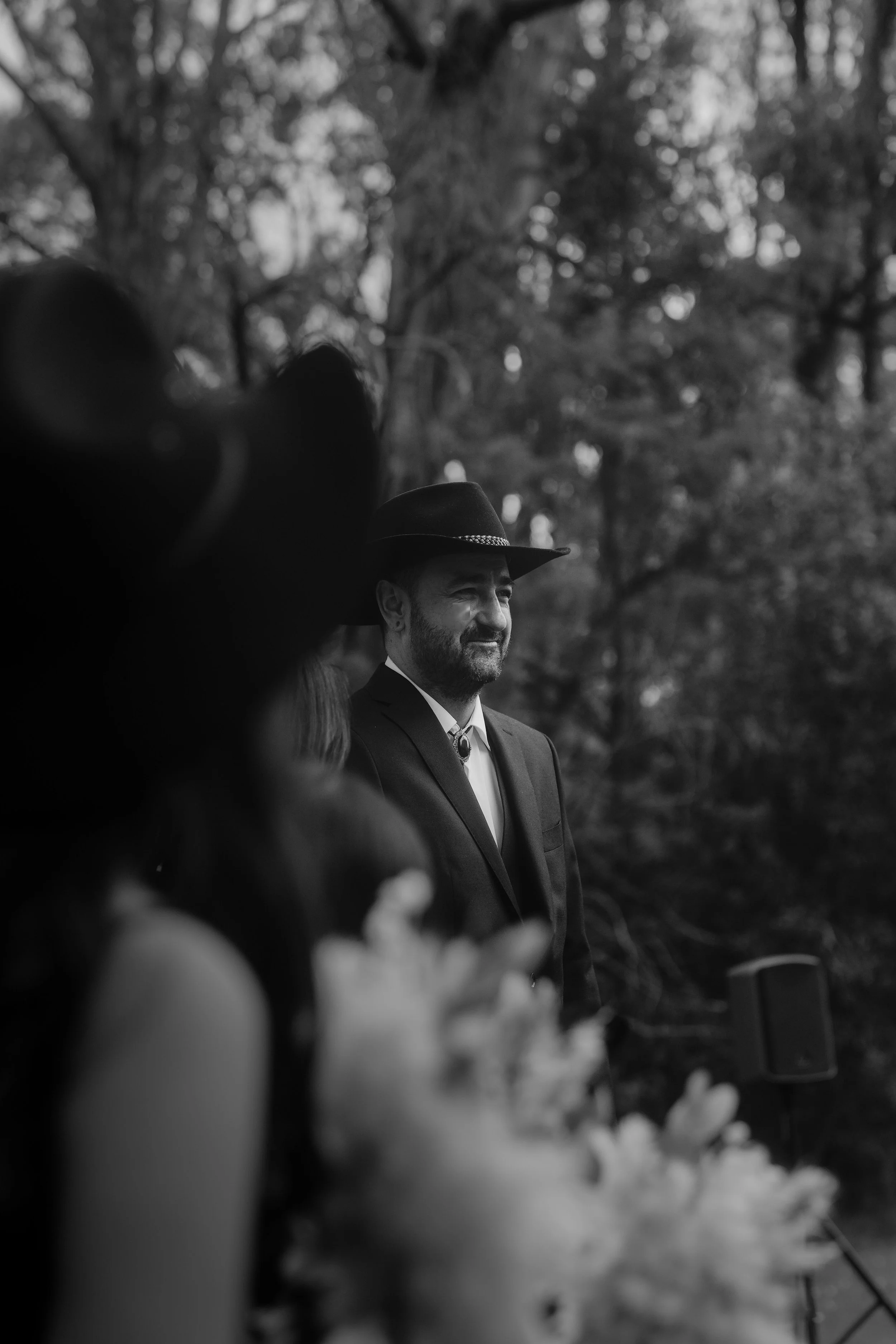 A black and white photo of a man with a beard wearing a suit and a wide-brimmed hat, standing outdoors with trees in the background, smiling slightly.
