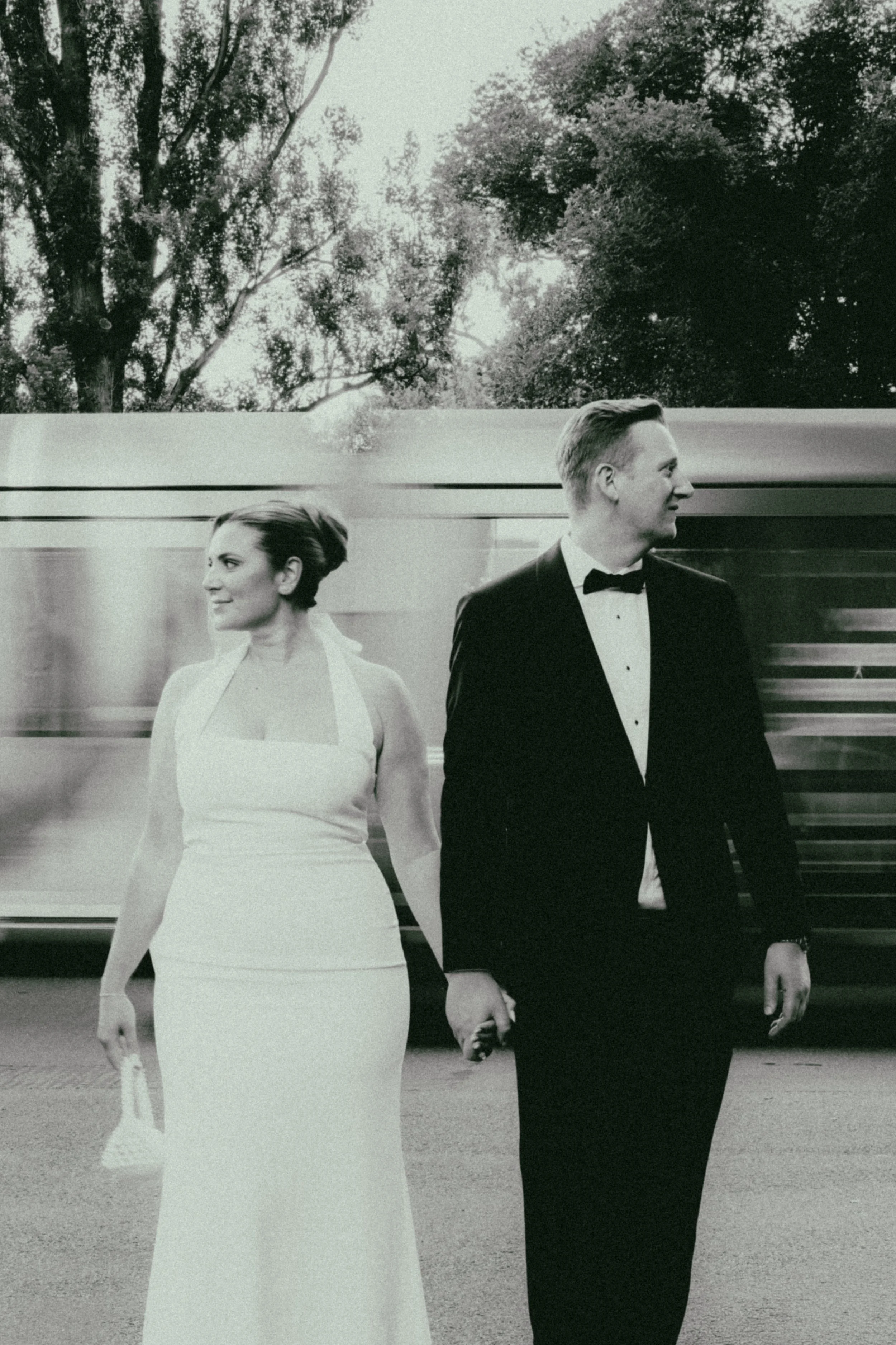 A black and white photo of a couple dressed in formal attire holding hands and walking outdoors, with a train passing behind them.