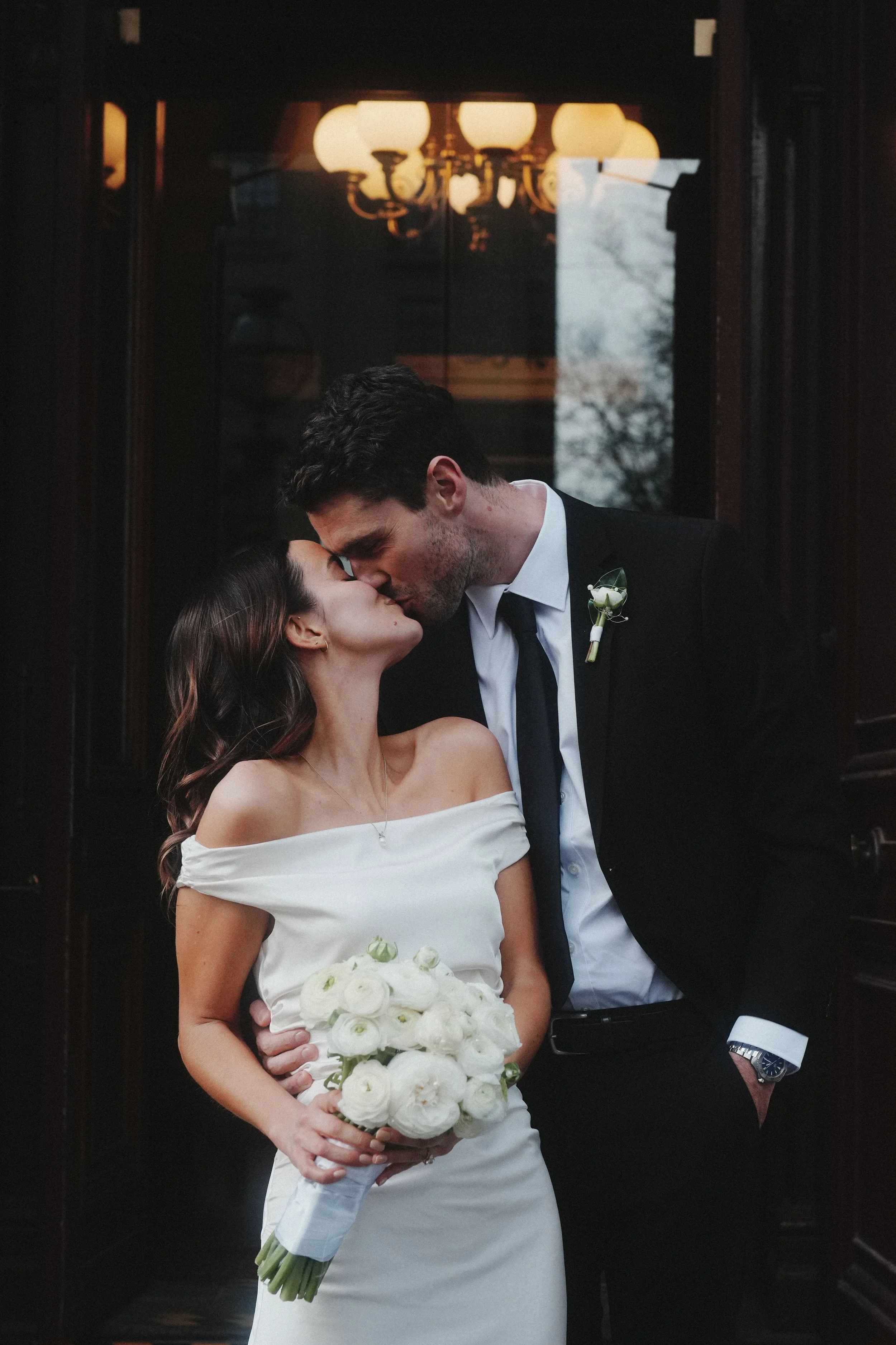 A bride and groom sharing a kiss indoors, with the bride holding a bouquet of white flowers and wearing an off-the-shoulder wedding dress, while the groom is dressed in a black suit and tie with a boutonniere, standing in front of dark wood paneling 