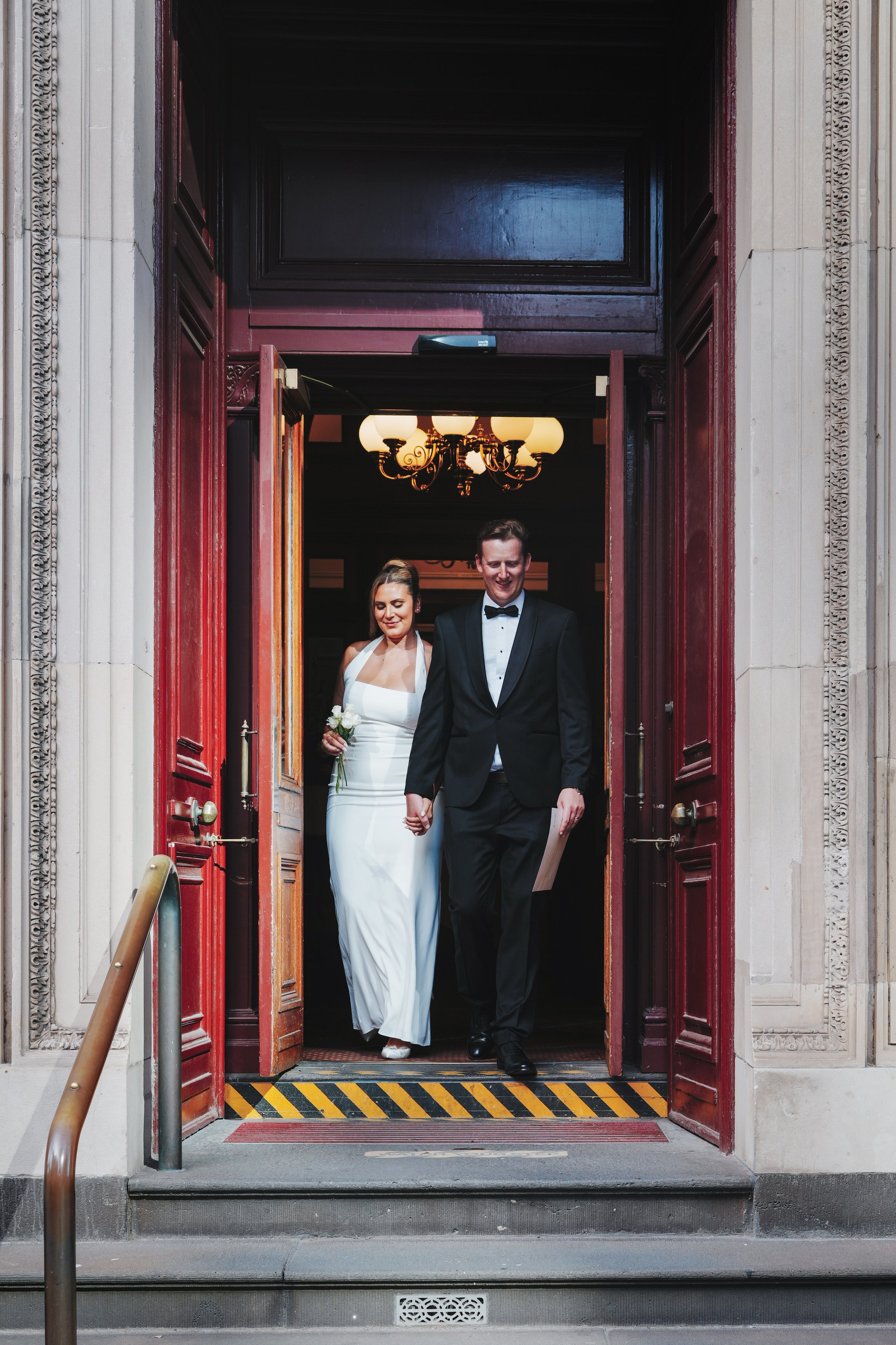 A newlywed couple walking out of a building holding hands. The bride is wearing a white wedding gown and holding a bouquet, and the groom is dressed in a black tuxedo. They are smiling as they exit the building through open red doors, with a chandeli
