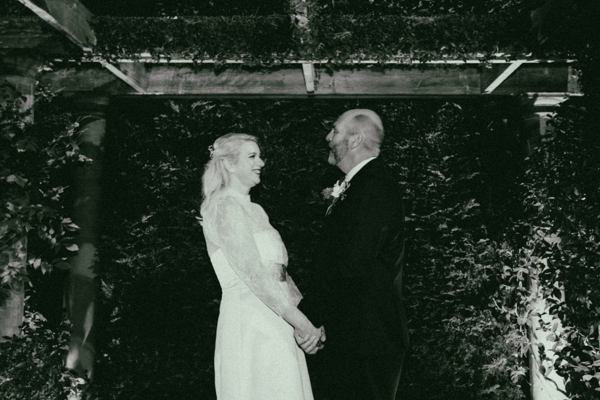 A bride and groom holding hands and smiling at each other during their wedding ceremony outdoors, surrounded by foliage.
