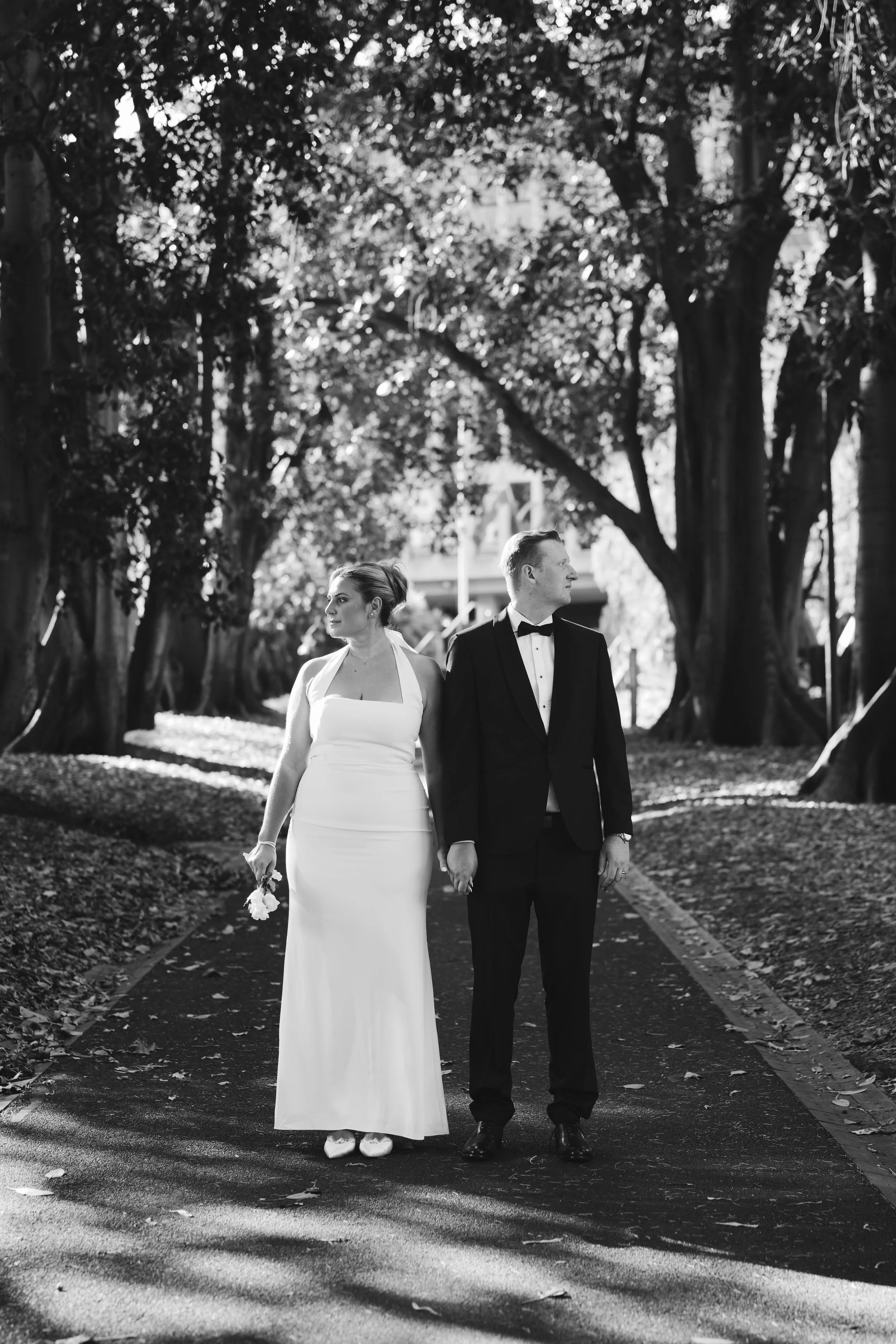 A bride and groom holding hands walking on a park path lined with trees, visible in black and white.