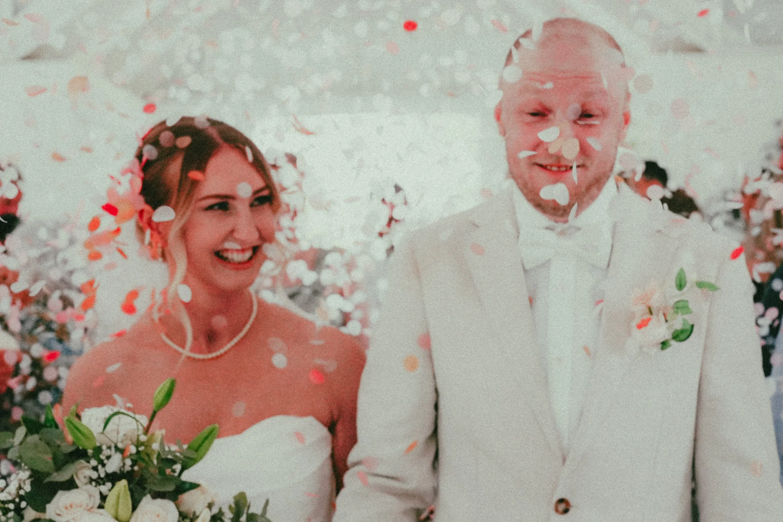 A bride and groom are smiling and celebrating with falling confetti at their wedding. The bride has blonde hair and is wearing a white dress, and the groom has short hair and is dressed in a white tuxedo. The scene is joyful and festive.