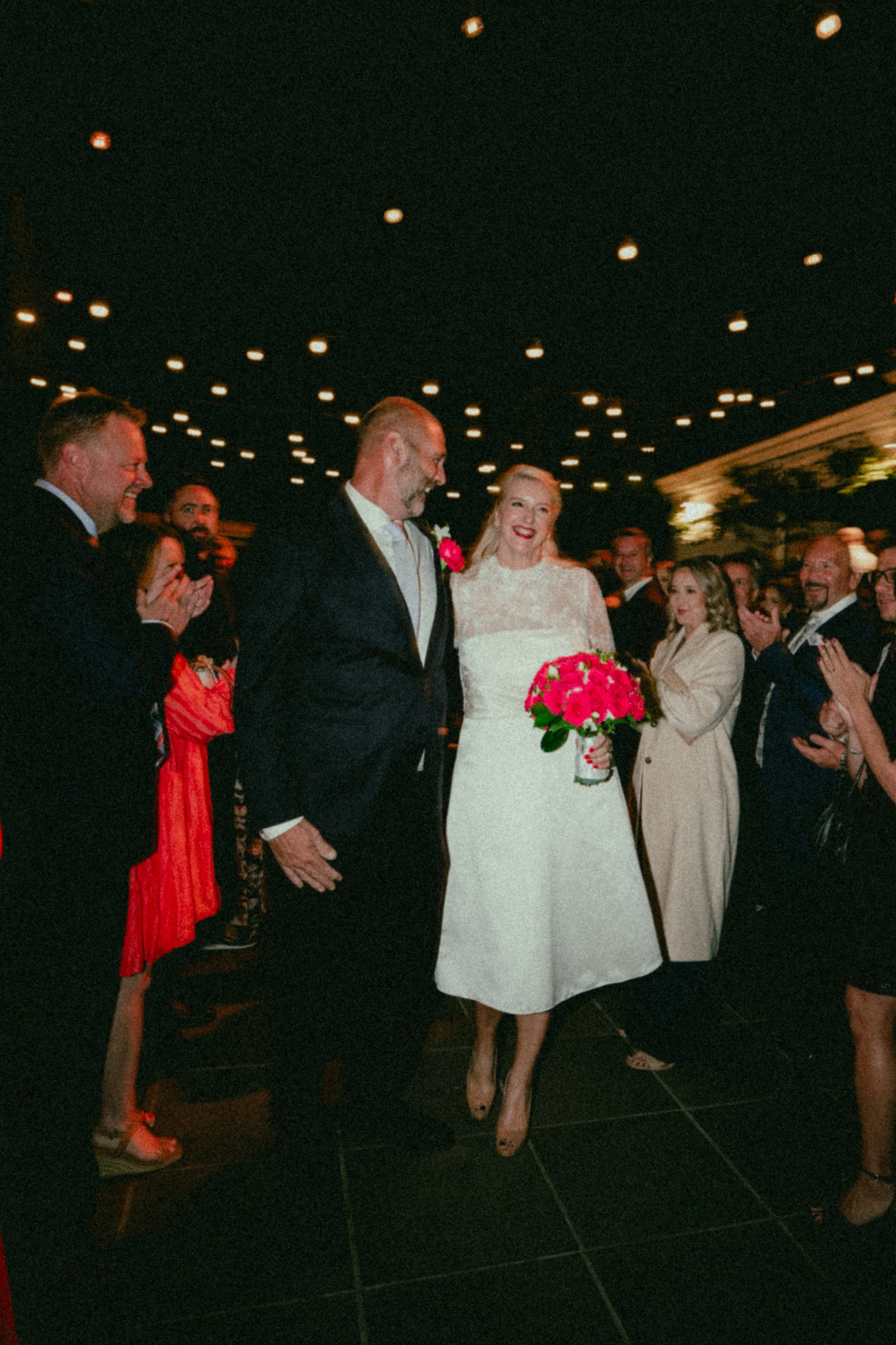 A wedding reception with a bride in a white lace dress holding a bouquet of pink flowers, standing next to a groom in a dark suit with a pink boutonniere, surrounded by smiling guests clapping and celebrating.