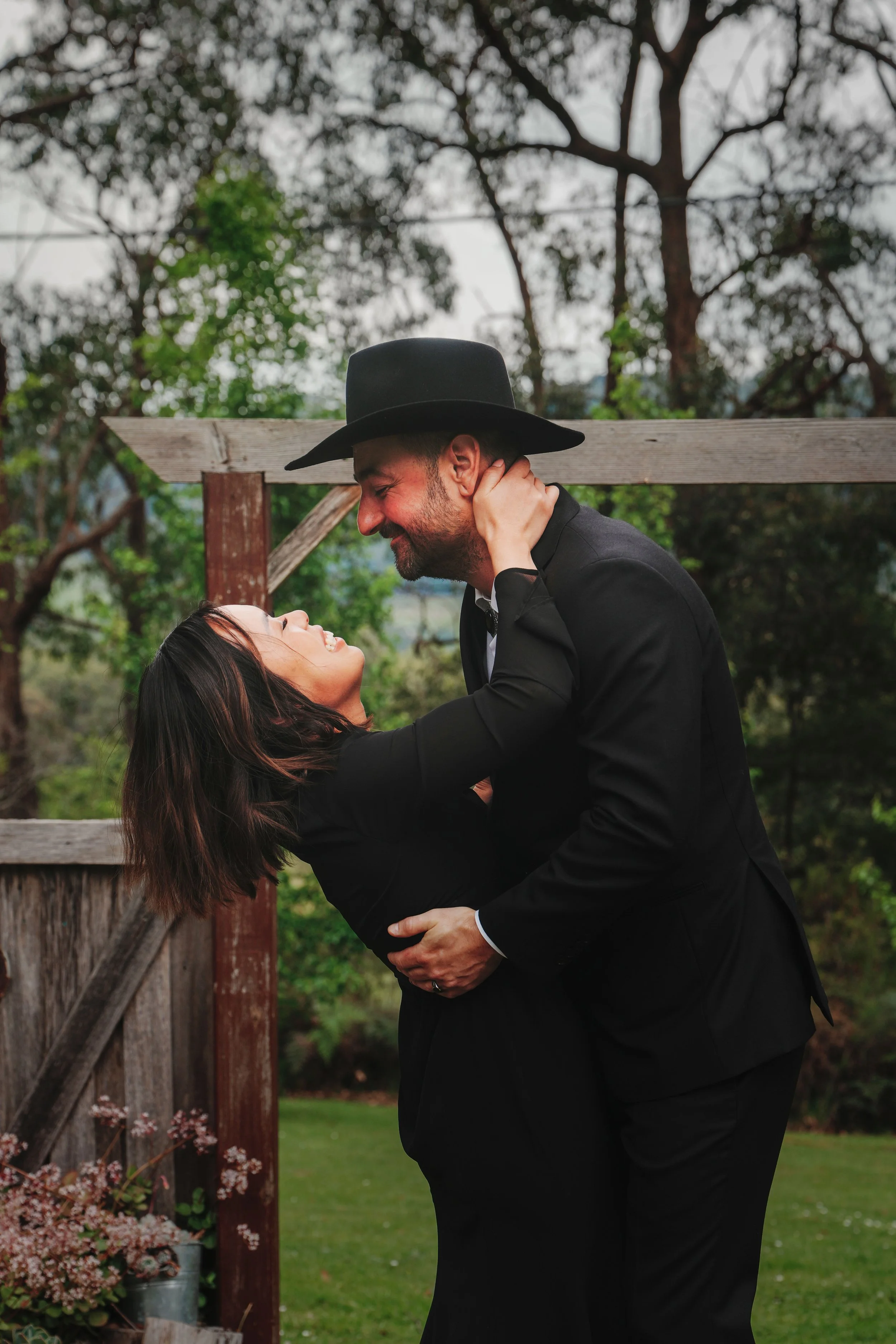 A man and woman sharing an intimate moment outdoors. The man, wearing a black suit and large black hat, gently holds the woman. She, with long dark hair, looks up at him with a smile. They are surrounded by trees and a wooden fence.