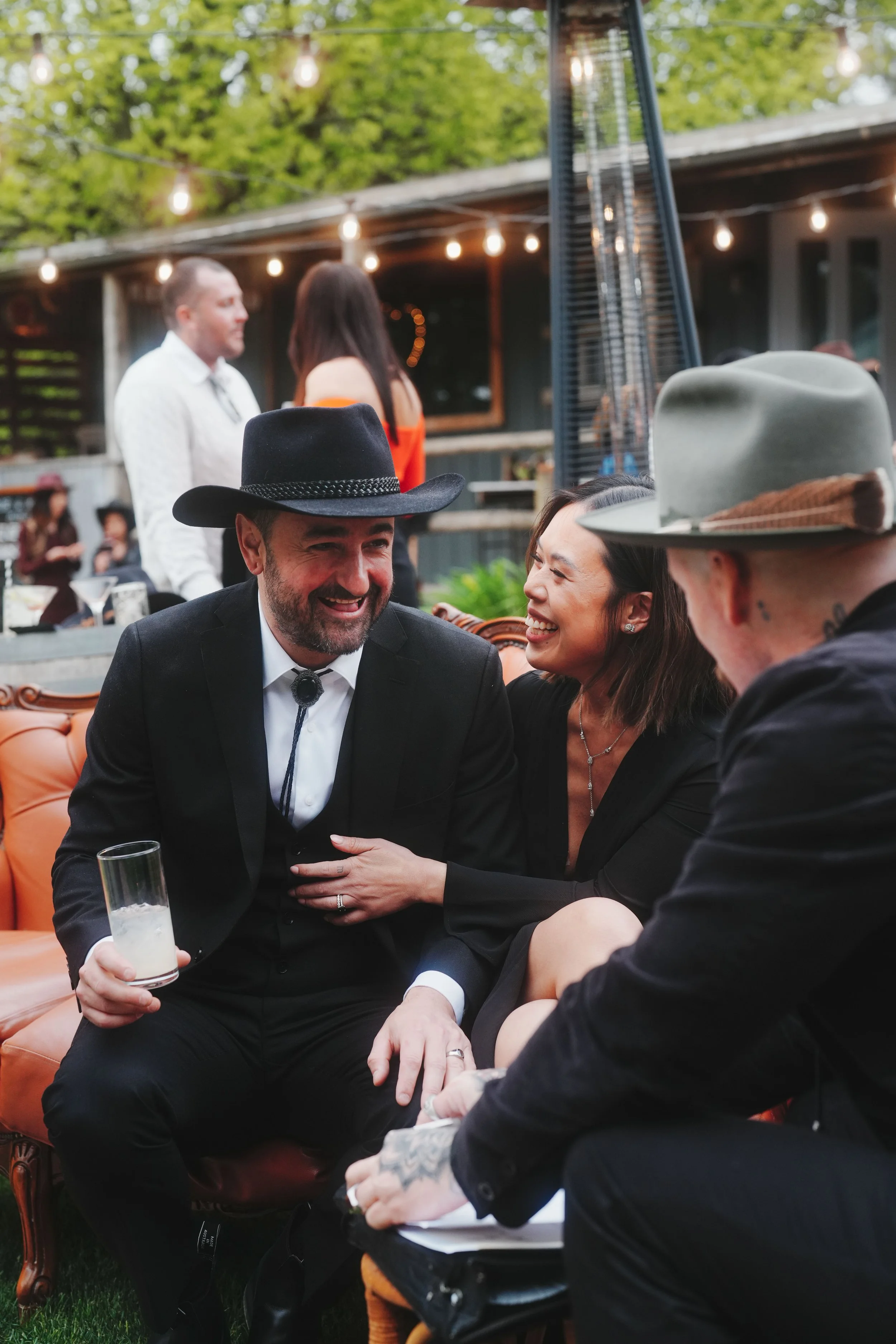 People enjoying an outdoor social gathering, talking, laughing, and sharing drinks, with string lights and trees in the background.