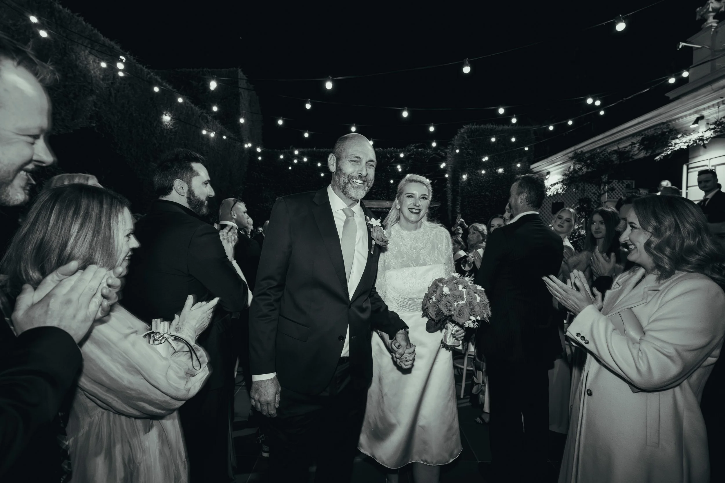 A black and white photo of a wedding reception with a bride and groom holding hands. The bride is holding a bouquet of roses, and both are smiling. Guests surround them, clapping and celebrating under string lights at night.
