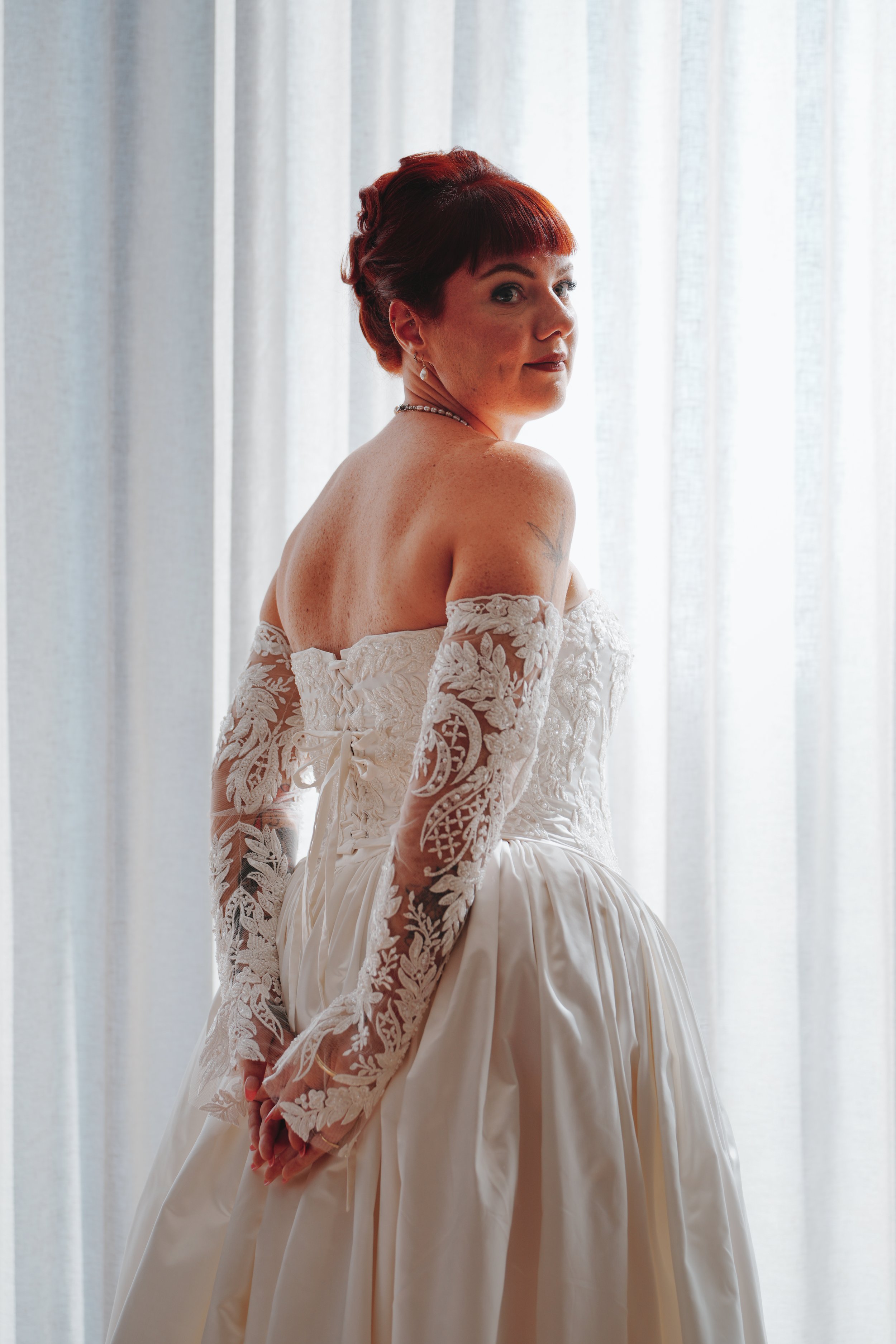 A woman with short red hair in a white lace off-shoulder dress standing in front of light-colored curtains, looking over her shoulder.