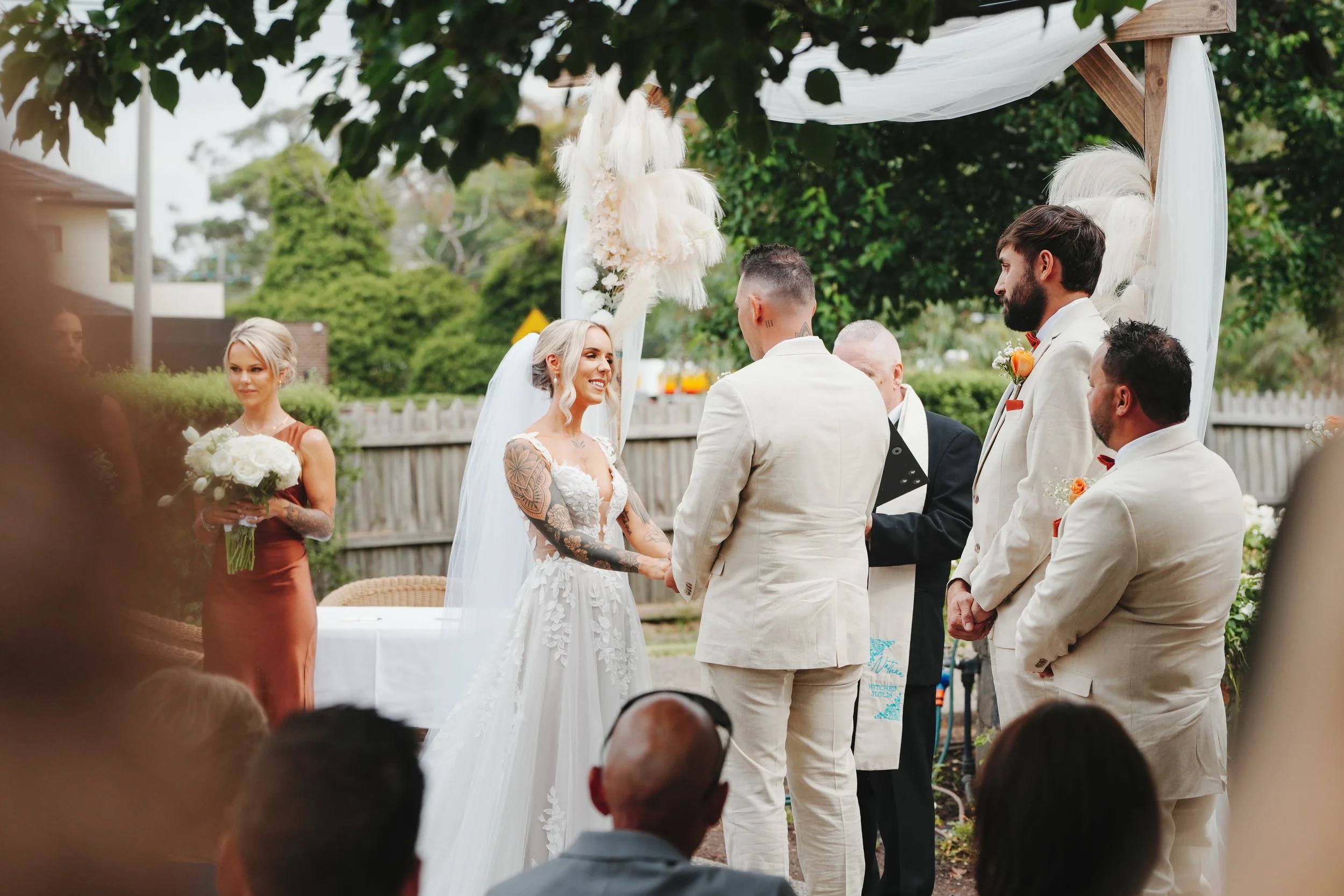 A wedding ceremony outdoors with a bride and groom holding hands, surrounded by friends and family. The bride wears a white lace wedding dress with tattoos on her arms, and the groom wears a white suit with a red bow tie. Bridesmaids in rust-colored 