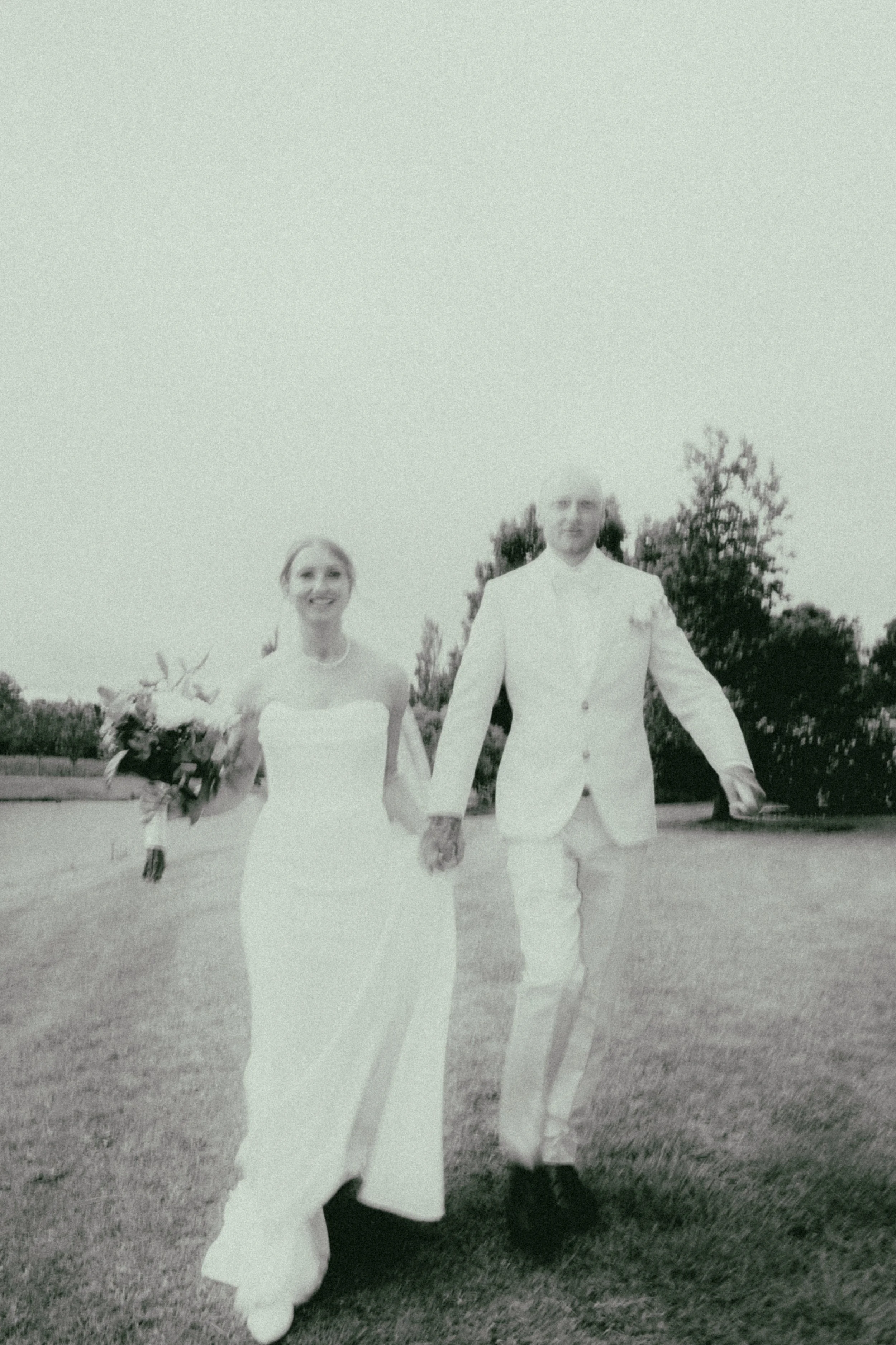A black-and-white photo of a man and a woman walking hand-in-hand outdoors on a grassy field, dressed in white formal clothes; the woman is holding a bouquet of flowers.