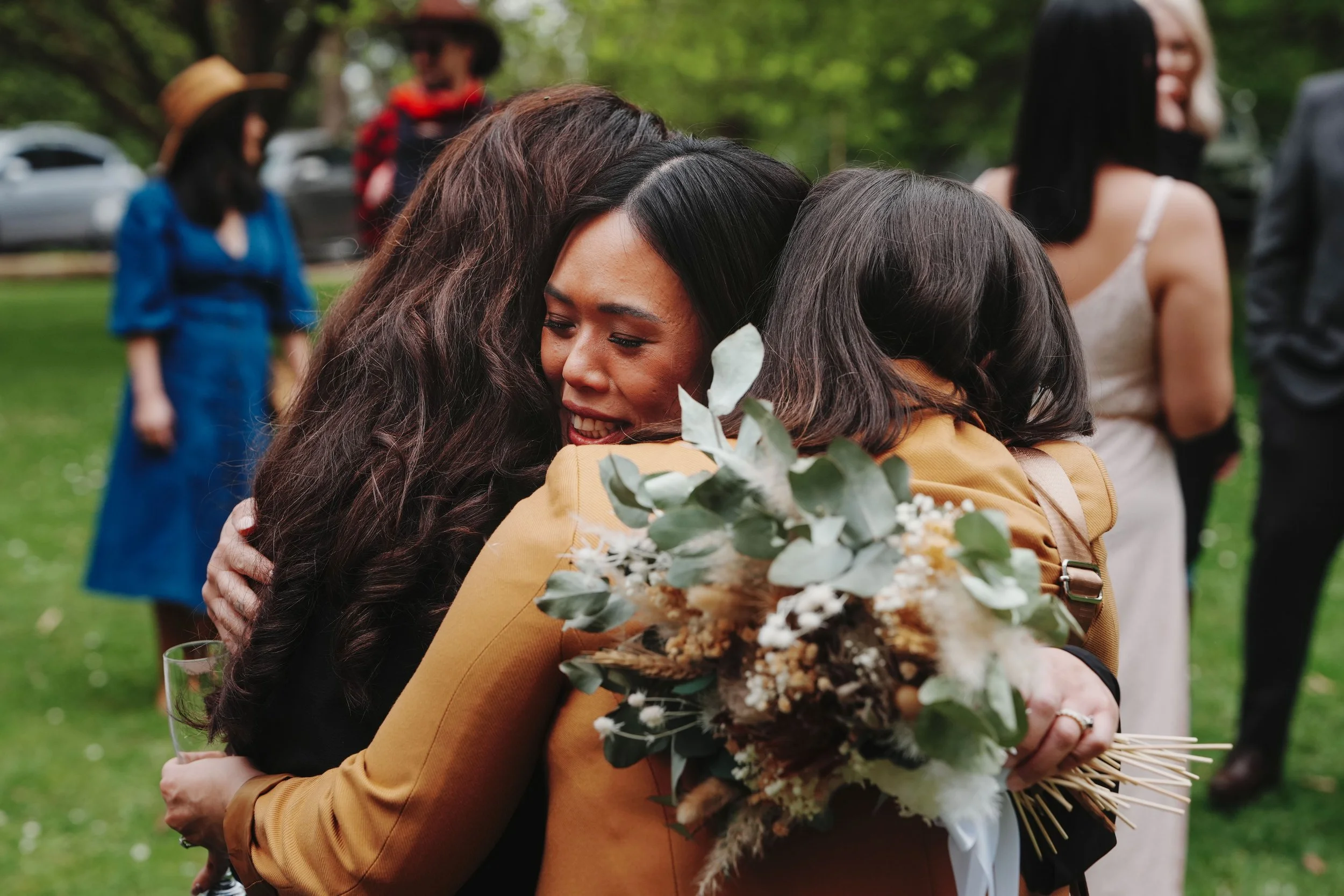 Two women embrace, one holding a bouquet of flowers and a glass of wine, at an outdoor gathering. Other people are visible in the background.